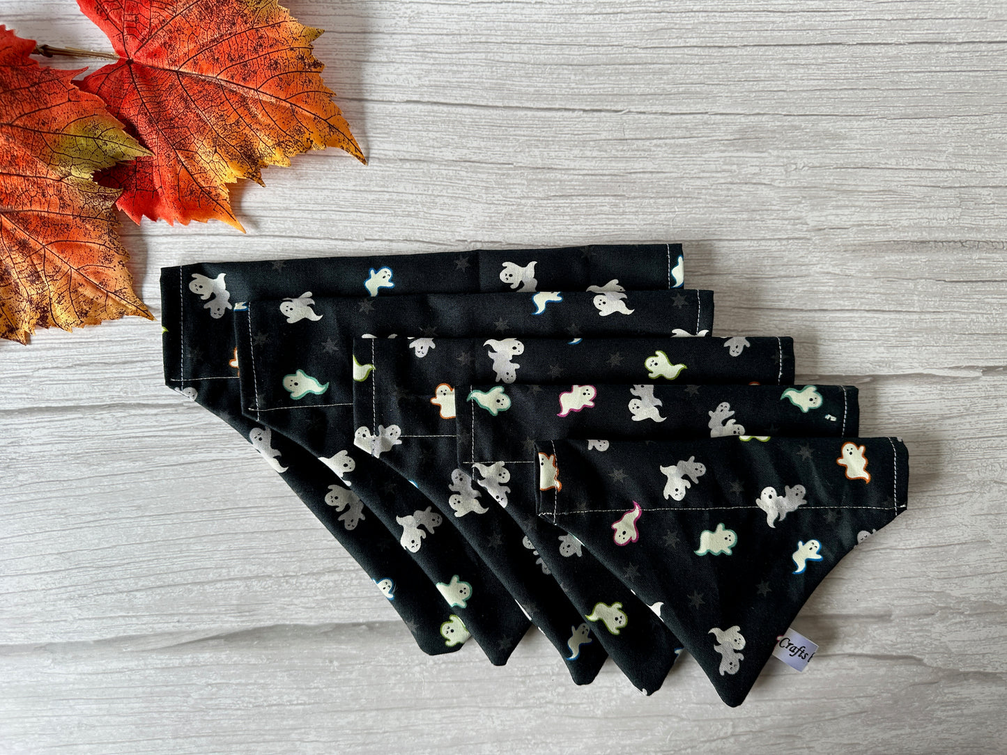 Six Crafts by Kate "Cotton Pet Bandana with collar attachment - Glow in the Dark Ghosts" are displayed on light wood. Each features small ghost patterns that glow in the dark. Two vibrant autumn leaves add a festive touch in the upper left corner.