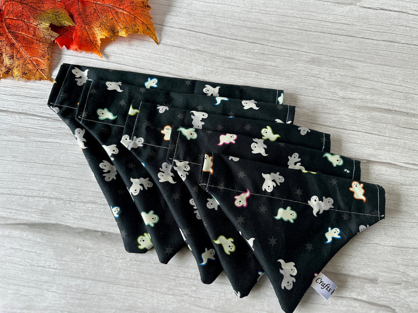 A Cotton Pet Bandana with collar attachment by Crafts by Kate, featuring a glow in the dark ghost pattern, is displayed on light wood. An artificial autumn leaf and a small "Craft" tag are also visible.