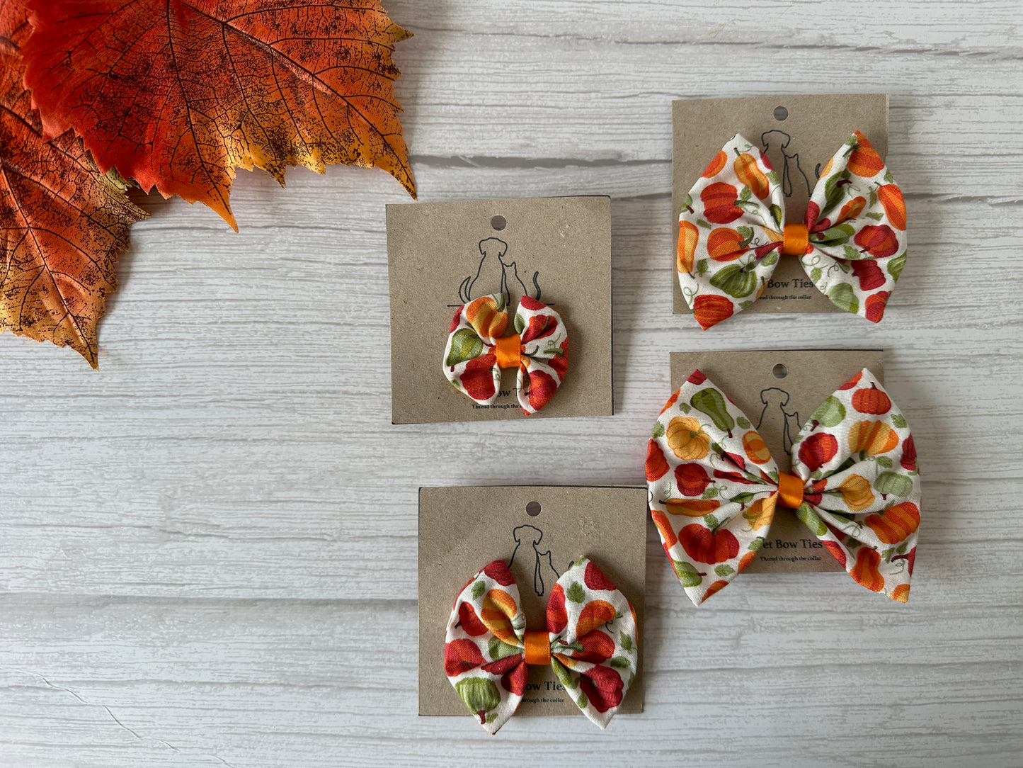 Three Cotton Pet Bows with collar attachments in Pumpkin Lover fabric by Crafts by Kate are displayed on brown cards atop light wood, with an orange autumn leaf in the upper left corner.