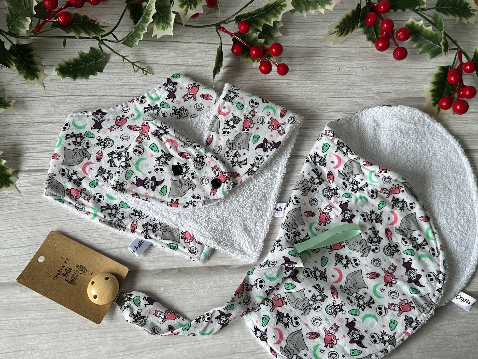 Flat lay of Crafts by Kate’s Cotton & Terry Towelling Baby Sets – Nightmare Before Christmas, featuring bandana bibs, dummy strap, and headband with festive prints. Background includes holly leaves and red berries on light wood.