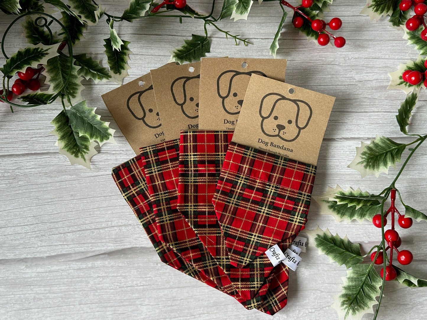 Four Crafts by Kate Cotton Pet Bandanas with collar attachment in Red Christmas Tartan are displayed on a white wooden surface, each featuring a brown dog-face tag and surrounded by festive green holly leaves with red berries.