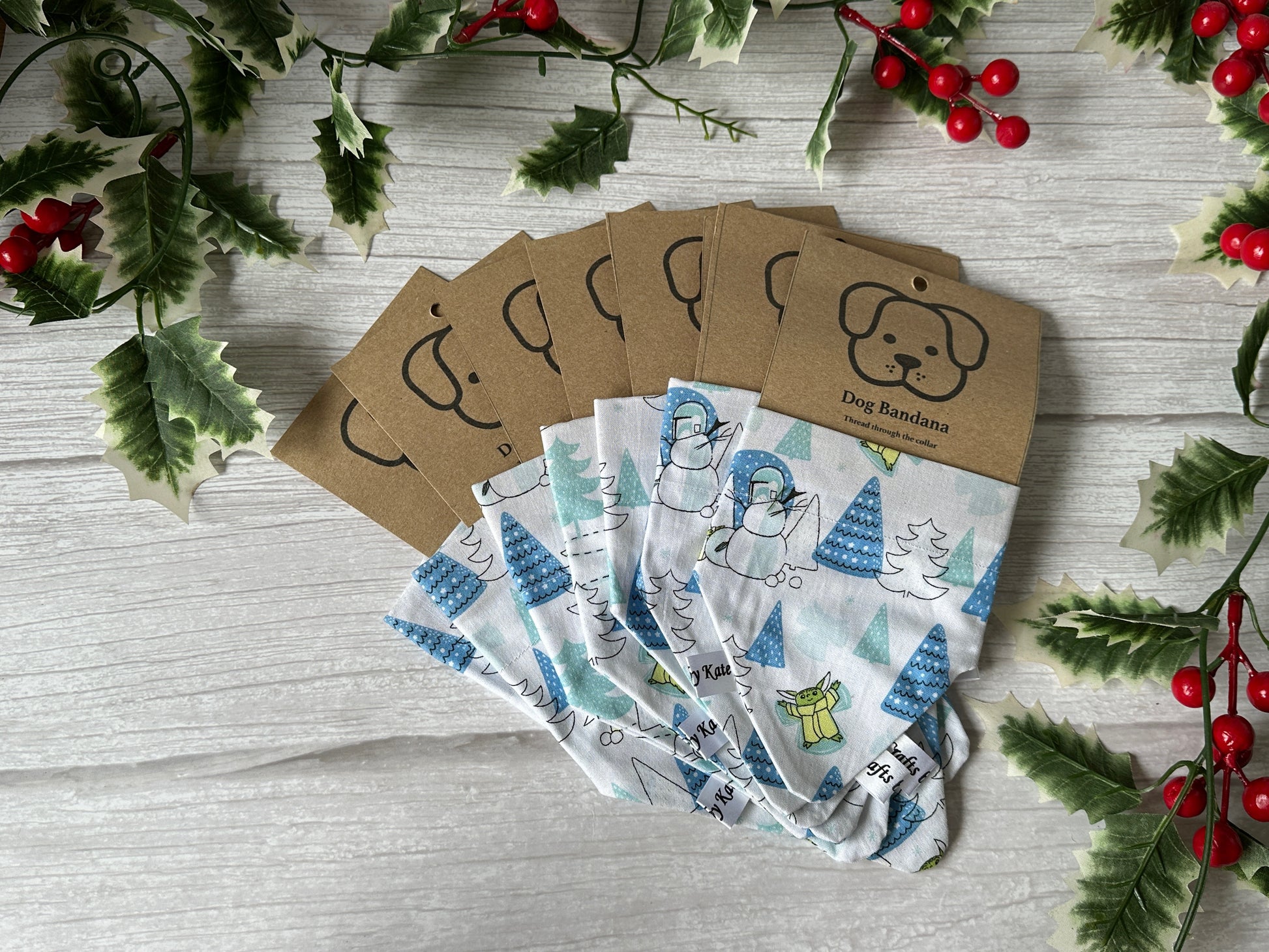 Five folded Cotton Pet Bandanas with collar attachment, featuring a Baby Yoda Christmas design from Crafts by Kate, are fanned out on a white wooden surface with holly and red berries, each packaged with brown labels showing a simple dog illustration.