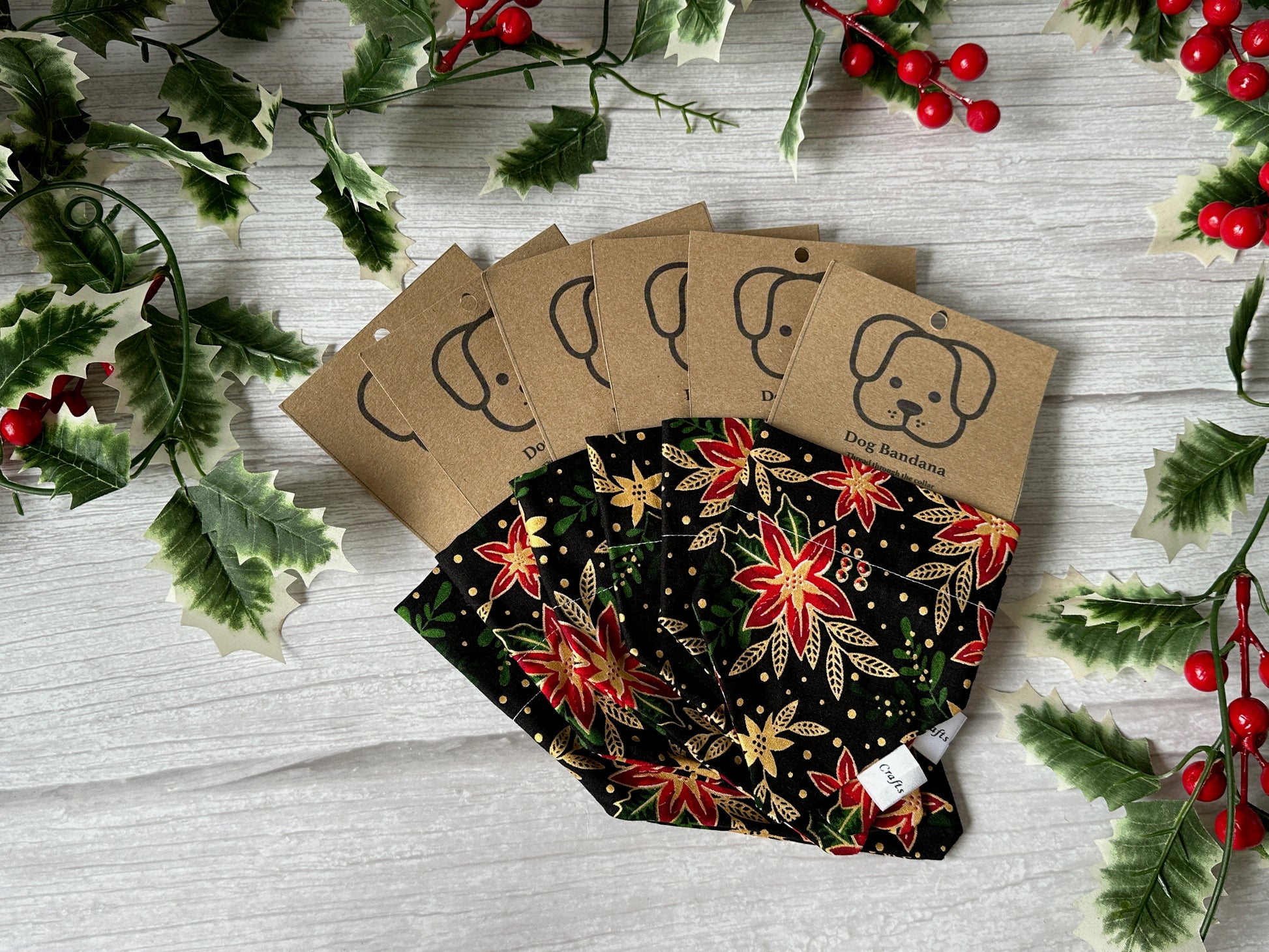 Five Crafts by Kate Cotton Pet Bandanas with collar attachment, featuring red, gold, and green poinsettia fabric, are displayed on a white wooden surface with holly leaves and berries for a festive holiday look.