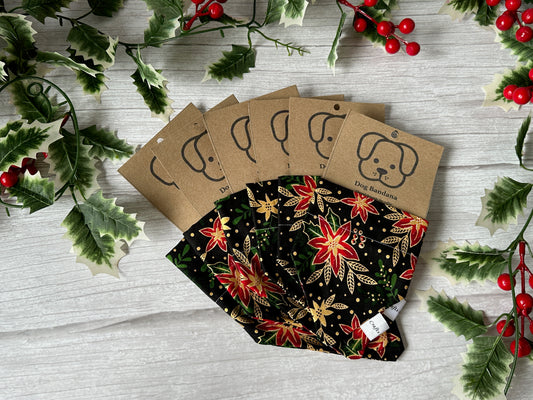 Five Crafts by Kate Cotton Pet Bandanas with collar attachment, featuring red, gold, and green poinsettia fabric, are displayed on a white wooden surface with holly leaves and berries for a festive holiday look.