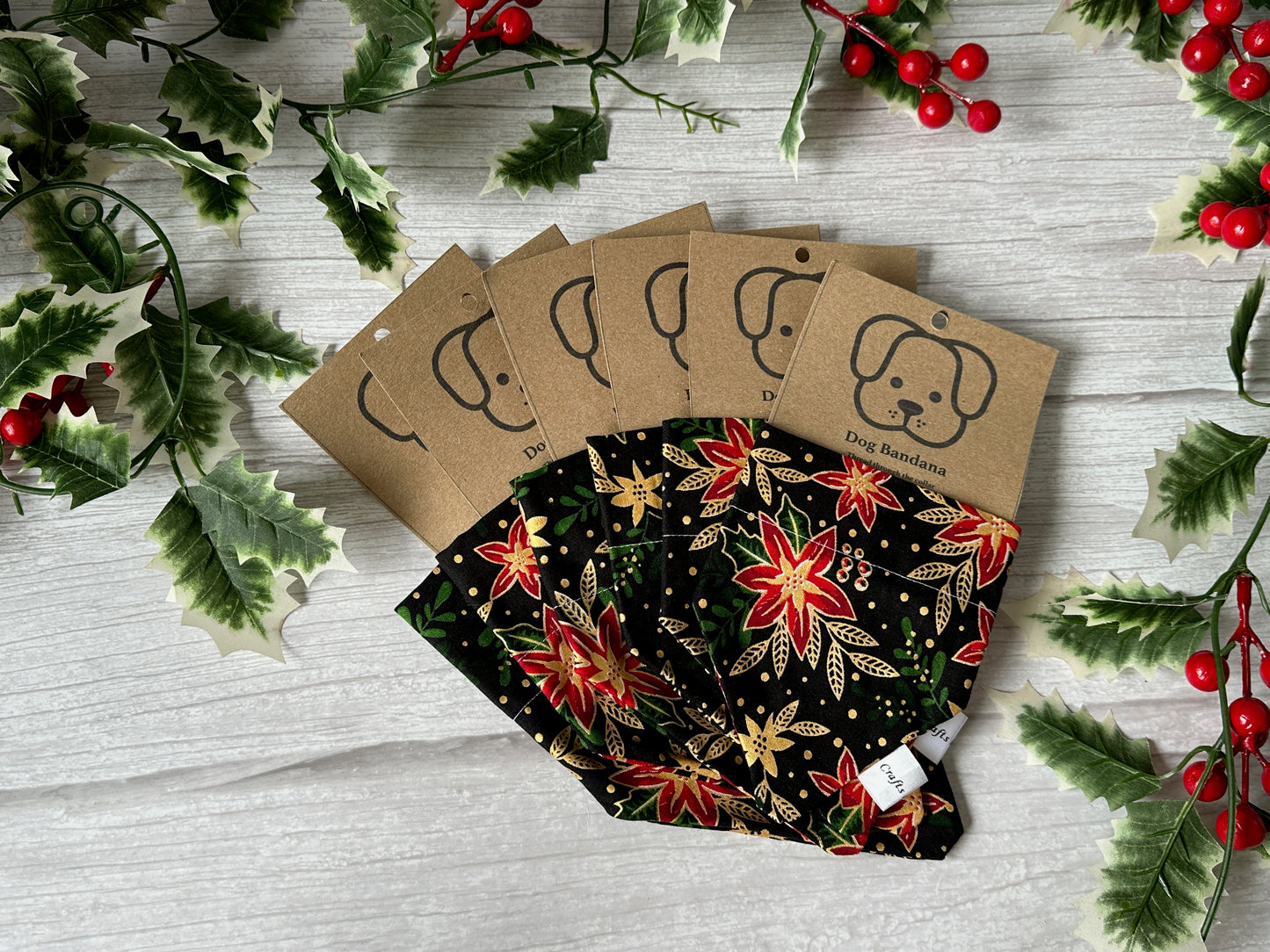 Five Crafts by Kate Cotton Pet Bandanas with collar attachment, in festive poinsettia prints, are displayed on a white wooden surface with faux holly and berries. Each is tagged with a dog illustration.