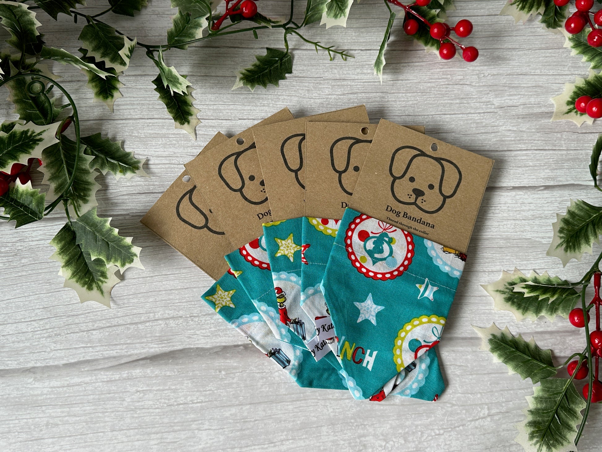 The Crafts by Kate Cotton Pet Bandana with collar attachment – Mr Grinch is shown on a light wooden surface, featuring festive holiday designs and a brown tag with a cartoon dog face. Holly and berries accent the image edges.