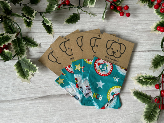 The Crafts by Kate Cotton Pet Bandana with collar attachment – Mr Grinch is shown on a light wooden surface, featuring festive holiday designs and a brown tag with a cartoon dog face. Holly and berries accent the image edges.