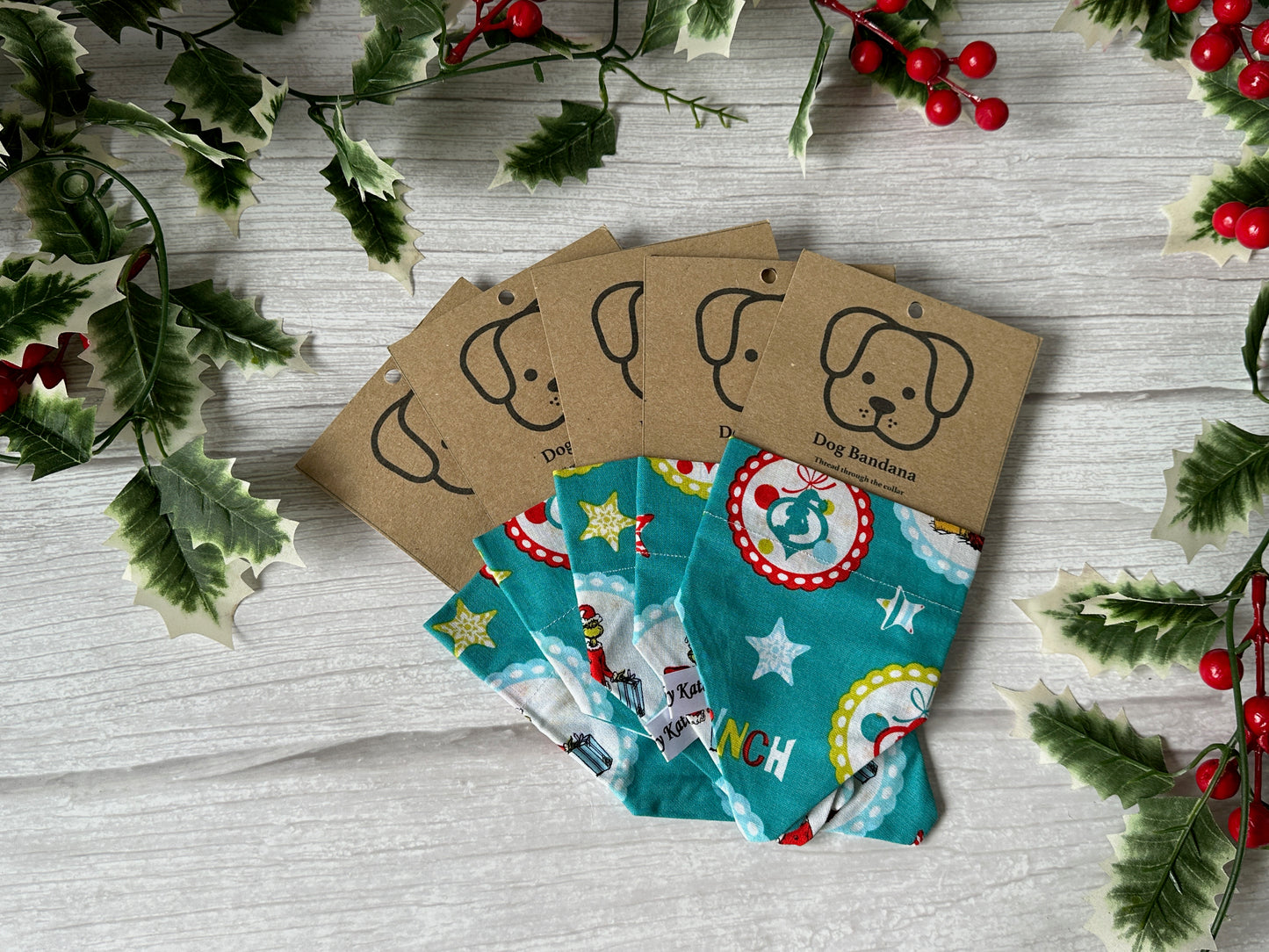 The "Cotton Pet Bandana with collar attachment - Mr Grinch" by Crafts by Kate is shown on a light wooden surface with holly and berries for a festive vibe. Each handcrafted bandana is packaged with a kraft label featuring a cute dog face illustration.
