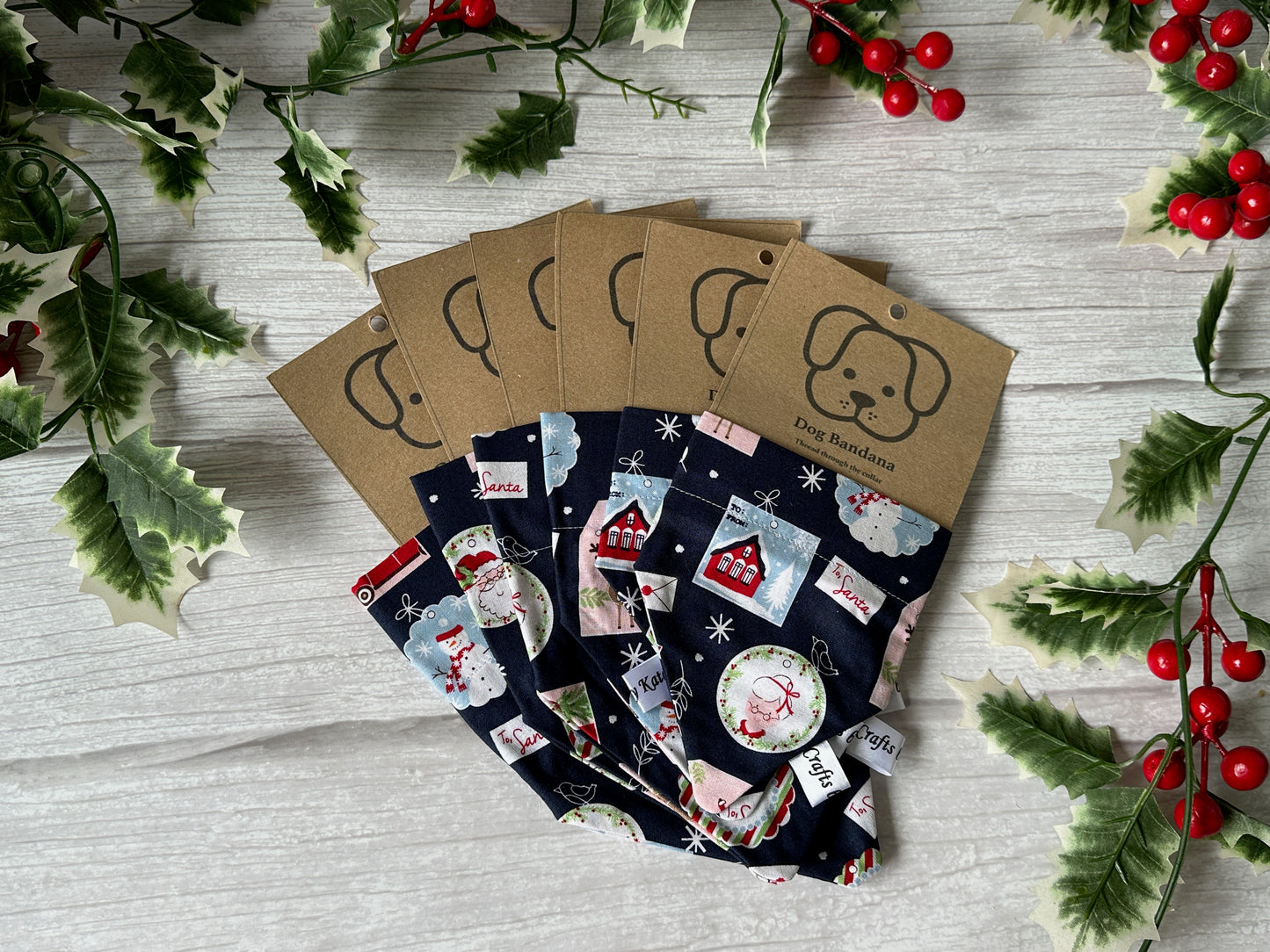 Six Cotton Pet Bandanas with collar attachments from Crafts by Kate, in festive Christmas Stamps fabric, are displayed on a light wooden surface with holly leaves and red berries. Each features a kraft tag with a dog face illustration.