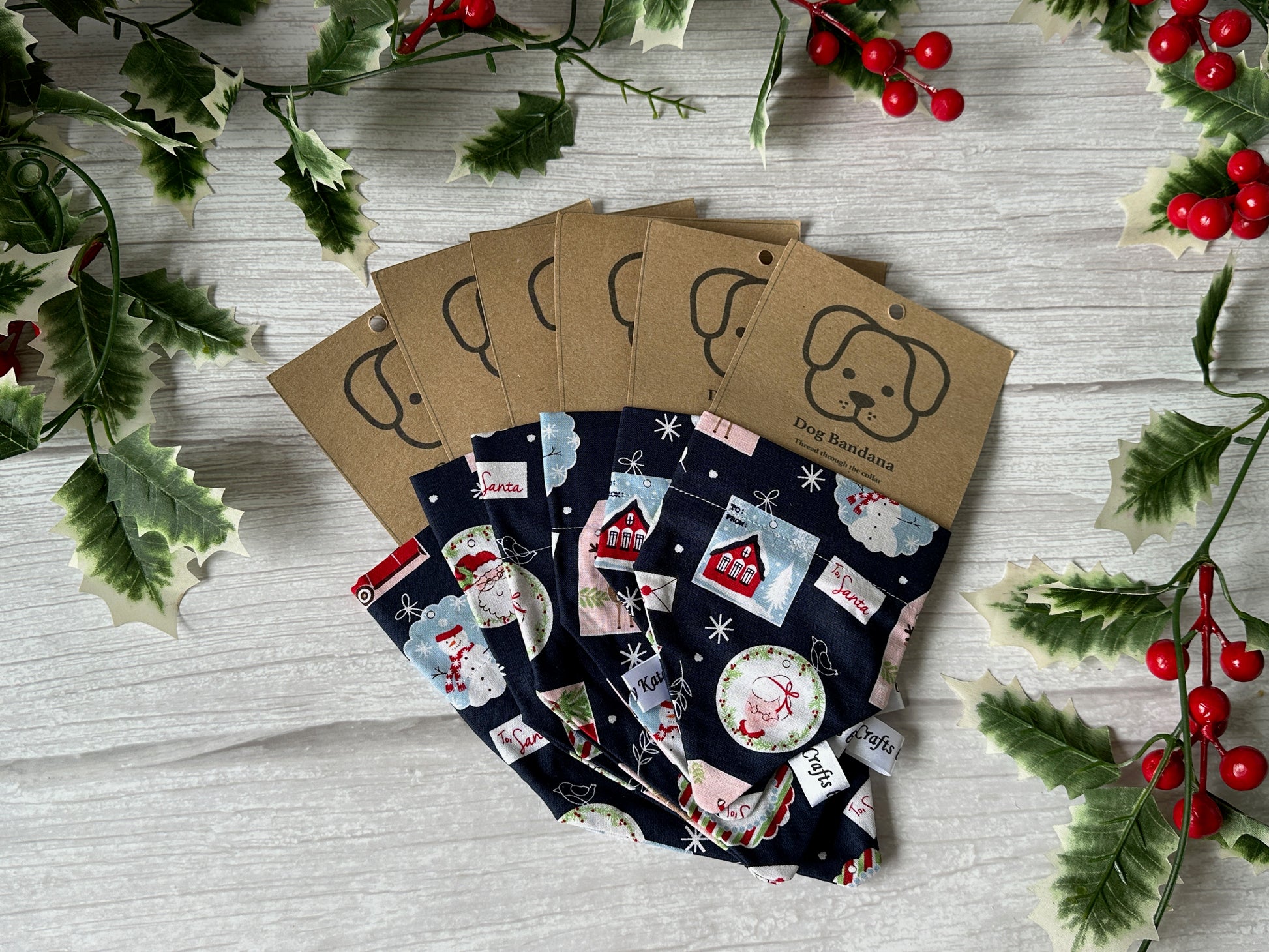 Six Cotton Pet Bandanas with collar attachments from Crafts by Kate, in festive Christmas Stamps fabric, are displayed on a light wooden surface with holly leaves and red berries. Each features a kraft tag with a dog face illustration.