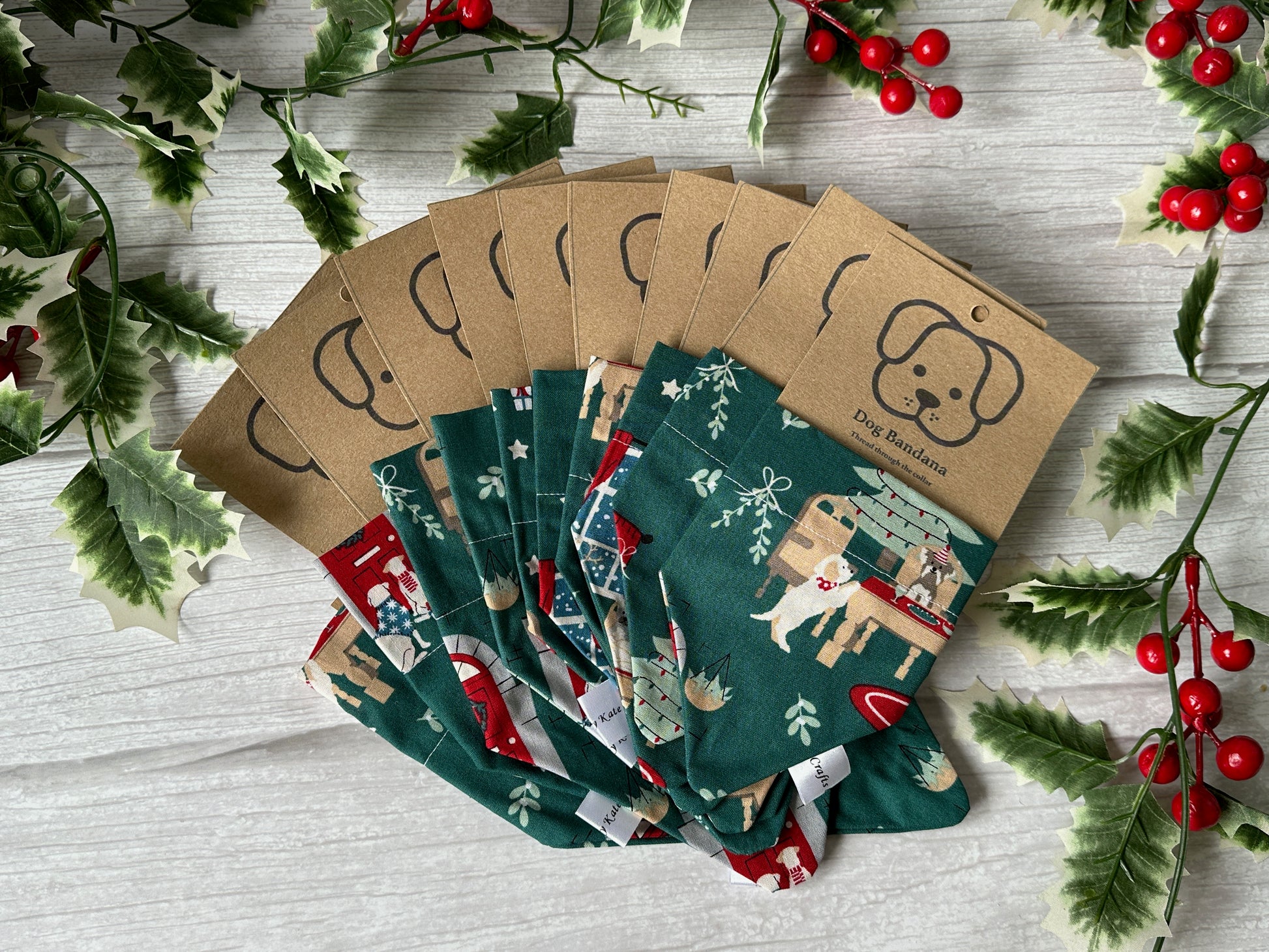 A fan display of Crafts by Kate Cotton Pet Bandana with collar attachment - A Dogs Christmas, featuring festive green patterns, each in branded brown cardboard packaging, surrounded by holly and red berries on a white wooden surface.