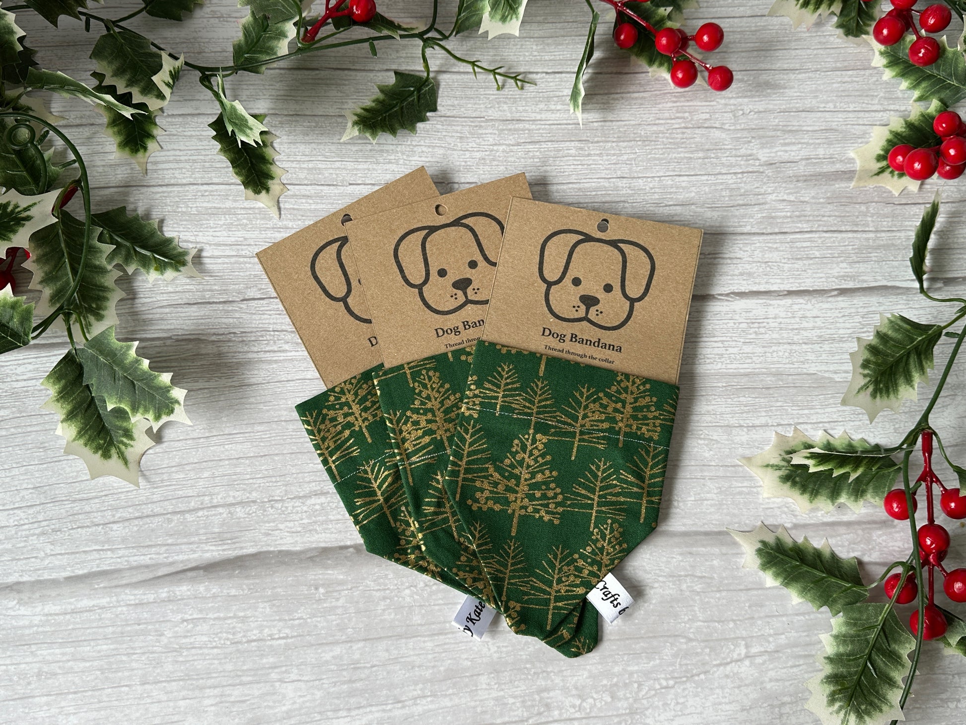 Three Cotton Pet Bandanas with collar attachment by Crafts by Kate, featuring green fabric with gold Christmas trees, are displayed on a light wooden surface amid holly leaves and red berries. Each has a brown tag with a dog face illustration.