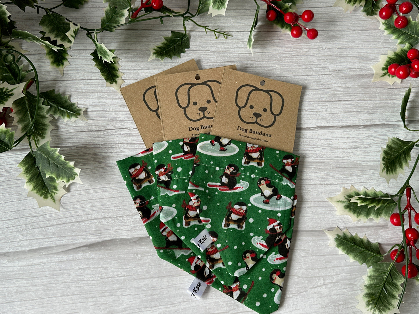 A Cotton Pet Bandana with collar attachment in a Skiing Penguins print by Crafts by Kate is displayed on a white wooden surface, decorated with holly leaves and red berries. Each has a kraft tag featuring a dog illustration and the words "Dog Bandana.