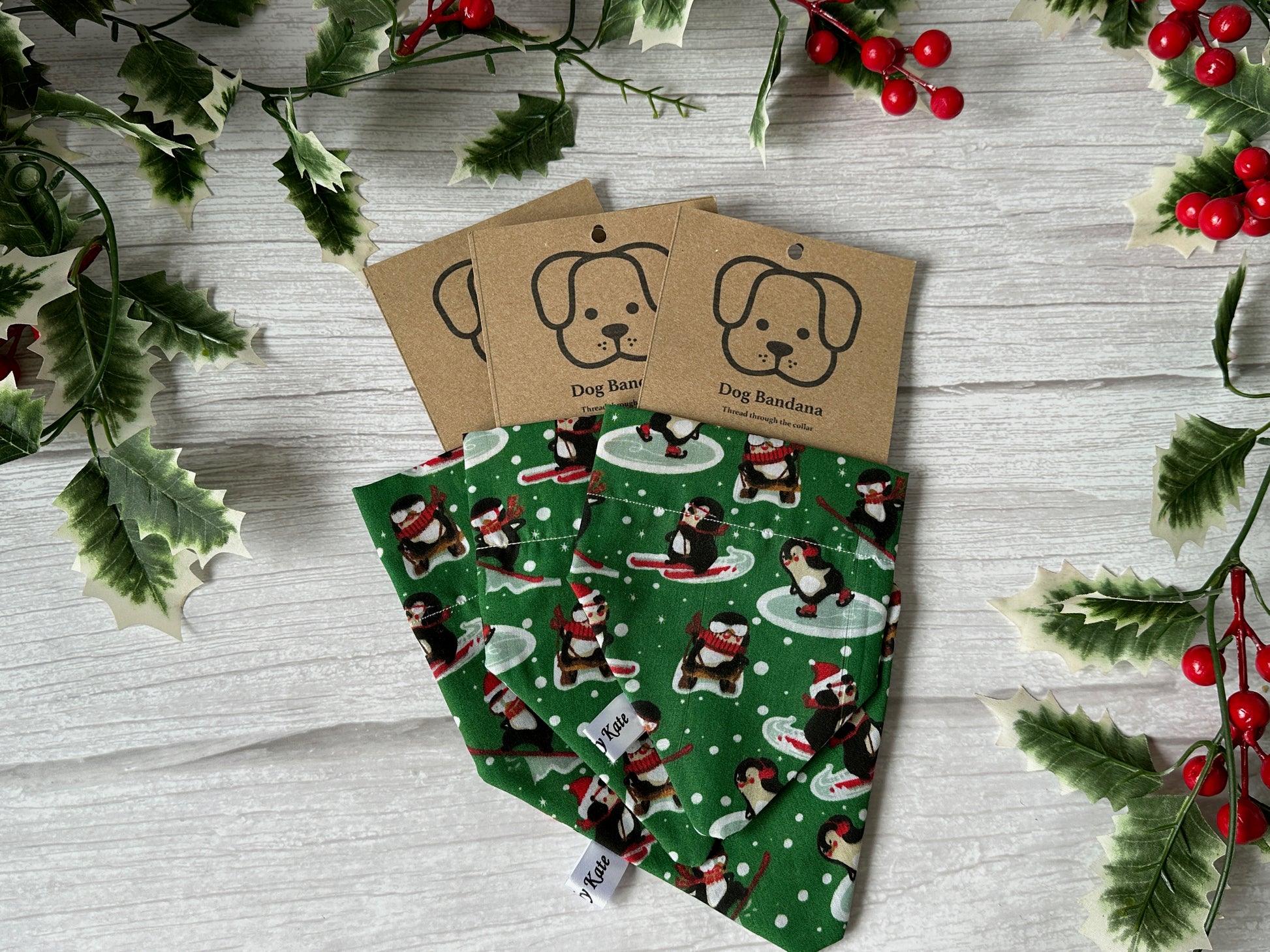 A Cotton Pet Bandana with collar attachment in a Skiing Penguins print by Crafts by Kate is displayed on a white wooden surface, decorated with holly leaves and red berries. Each has a kraft tag featuring a dog illustration and the words "Dog Bandana.
