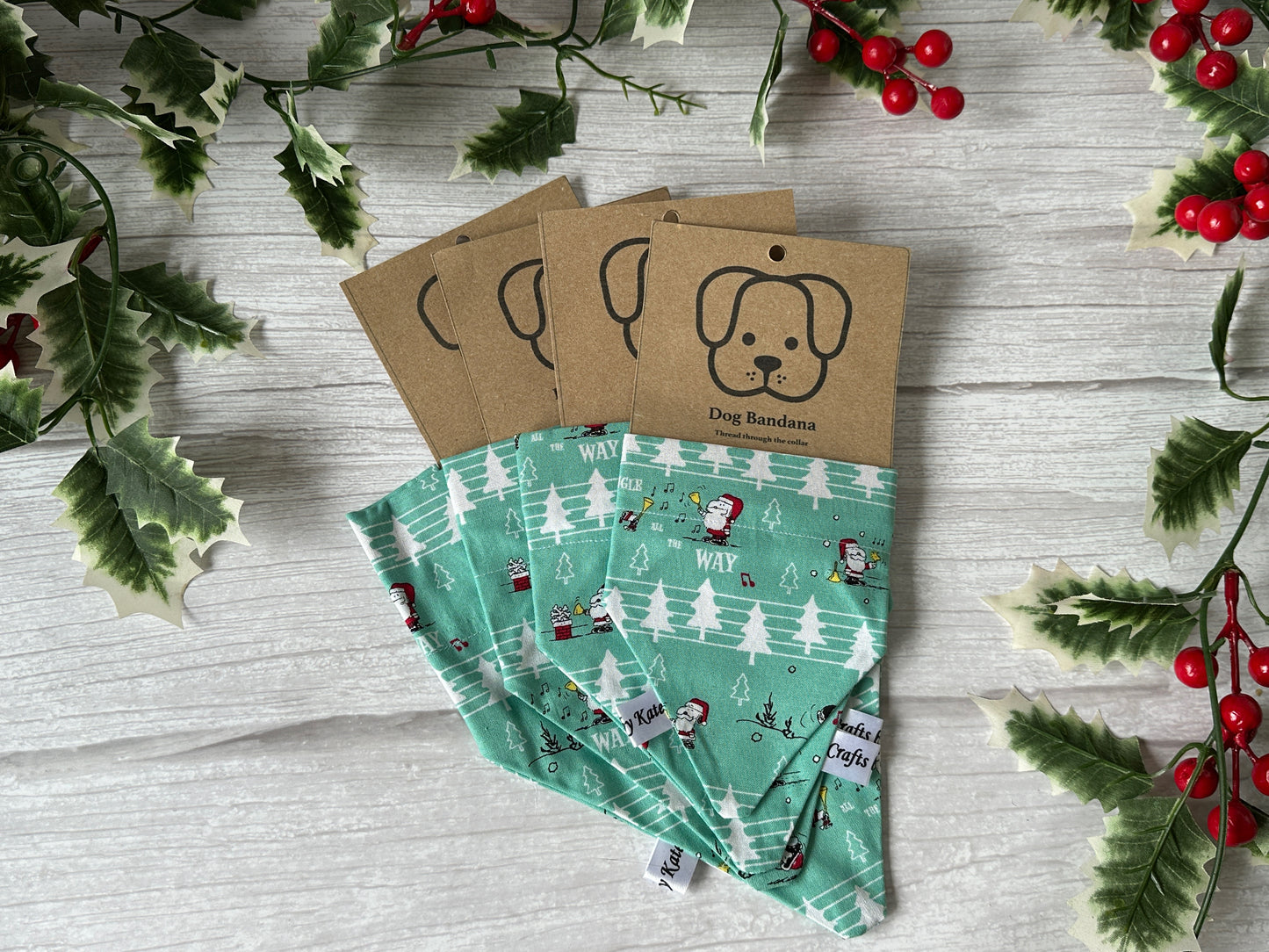 Three green "Cotton Pet Bandana with collar attachment - Snoopy Christmas" by Crafts by Kate, each with winter patterns and cardboard tags, arranged on a white wooden surface with festive holly decor. Perfect for holiday celebrations.