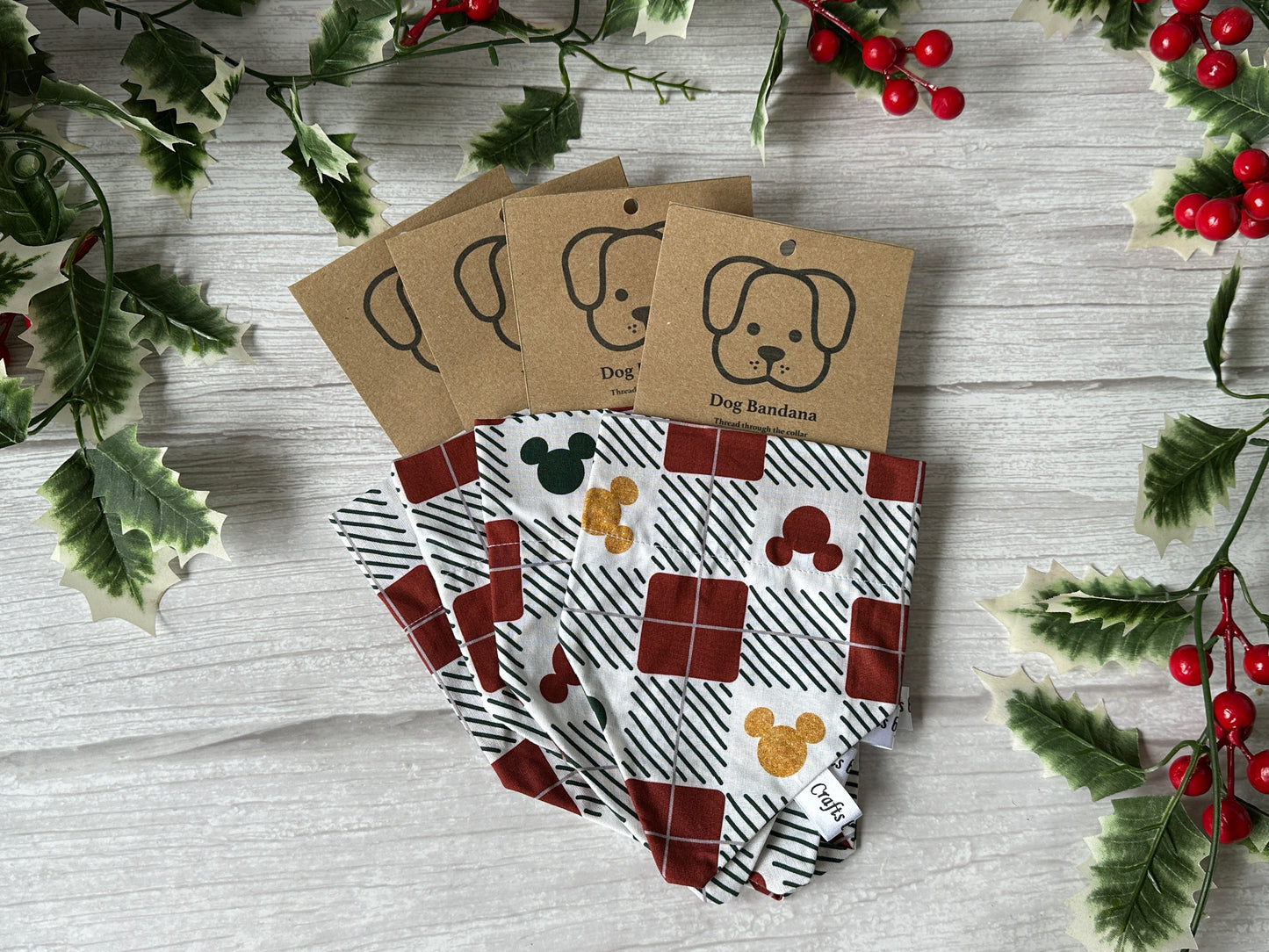 Four Crafts by Kate Cotton Pet Bandanas with collar attachments in Micky Mouse Tartan Christmas patterns are displayed on wood, surrounded by holly and berries. Each bandana features a brown label with a cartoon dog face—great for festive pets!.