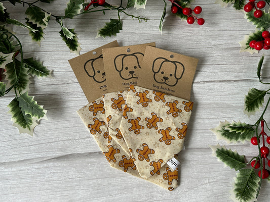 Three Cotton Pet Bandanas with collar attachments, Gingerbread Lovers by Crafts by Kate, are displayed folded on a light wooden surface with holly leaves and red berries. Each handcrafted bandana has a brown tag featuring a dog illustration.