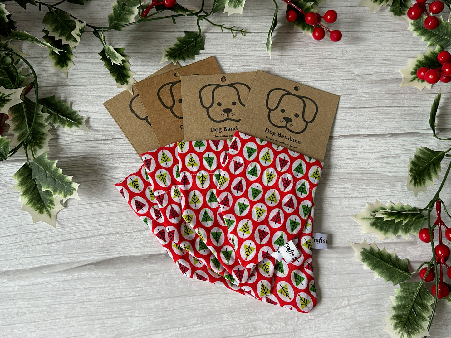 Three "Crafts by Kate" Cotton Pet Bandanas with collar attachments, featuring red, green, and white Christmas tree patterns, are displayed on a light wood surface with holly and red berries.