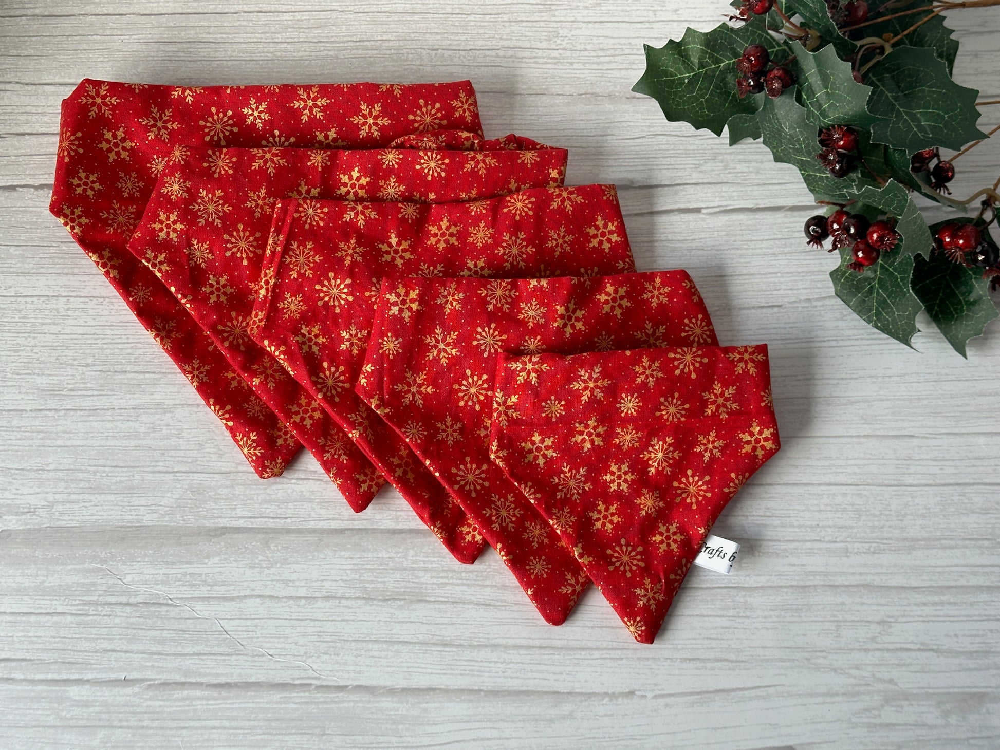 Five Cotton Pet Scrunchie Bandanas in Red & Gold Snowflakes by Crafts by Kate are stacked on light wood beside artificial holly, making the perfect festive pet accessory.