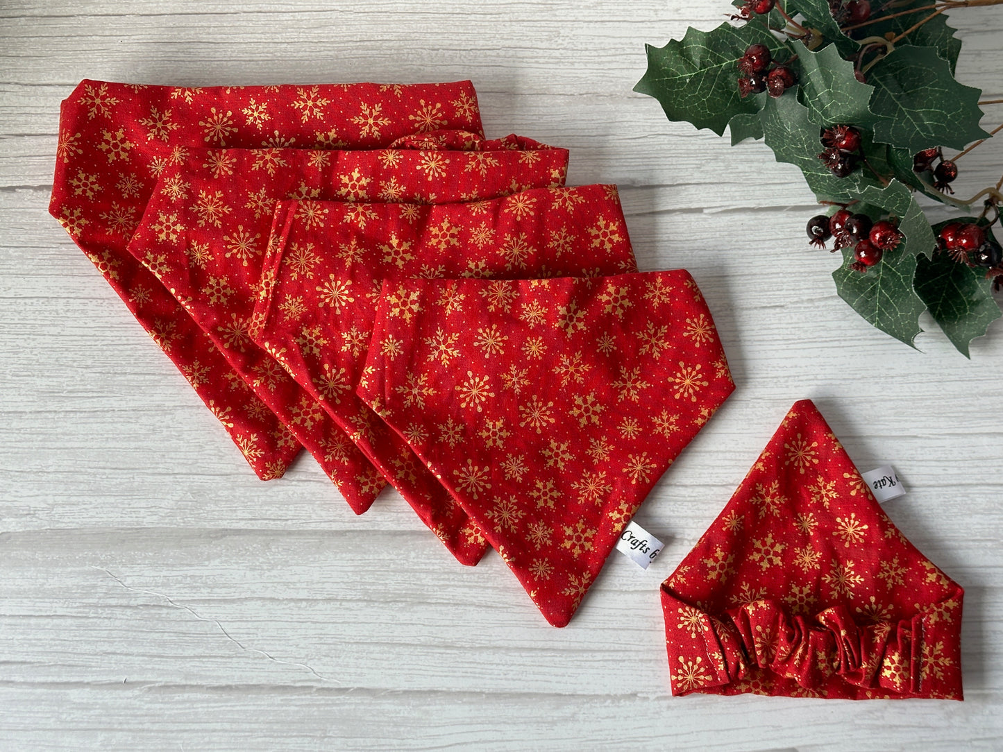 Five Crafts by Kate “Cotton Pet Scrunchie Bandana - Red & Gold Snowflakes” are displayed on light wood, with a matching cotton pet scrunchie and an artificial holly decoration featuring berries arranged beside them.