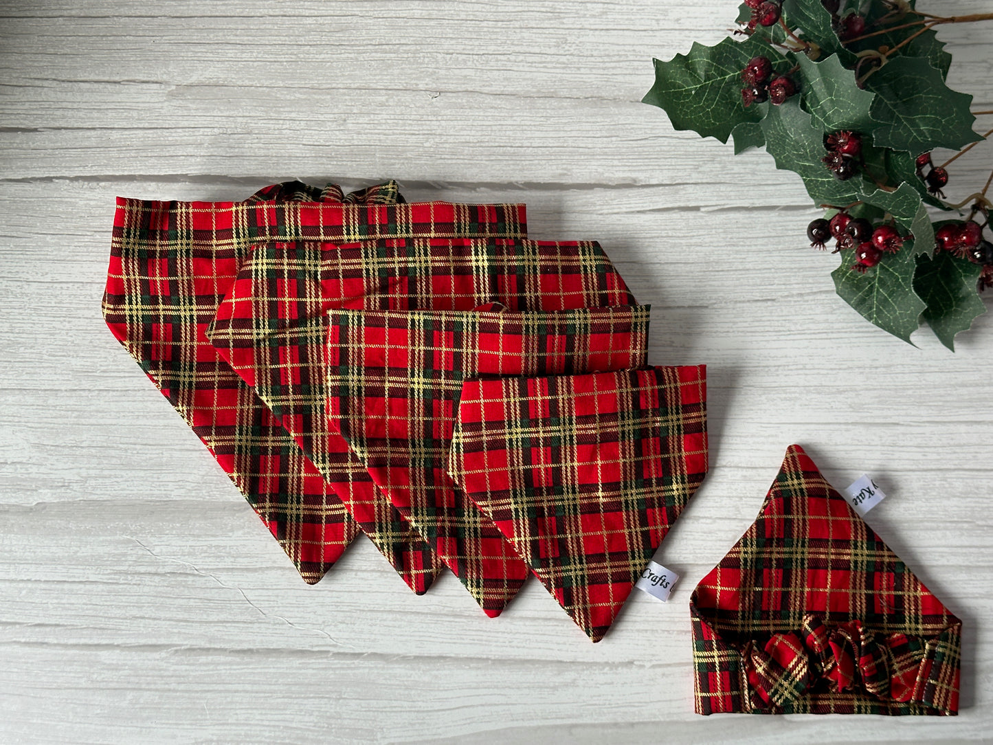 Crafts by Kate’s Cotton Pet Scrunchie Bandana in Red & Gold Tartan is displayed with other plaid styles, arranged by size on a light wood surface, accented with holly and berries in the top right corner.