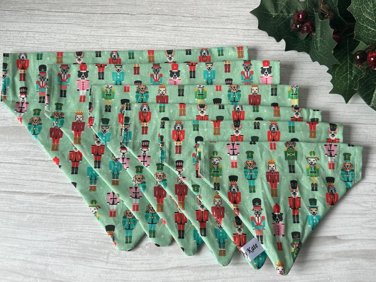 Five green cloth napkins with a nutcracker soldier pattern are fanned out on light wood. Holly and berries in the corner echo the festive spirit of the Crafts by Kate Cotton Pet Bandana with collar attachment—Puppy Nutcracker.