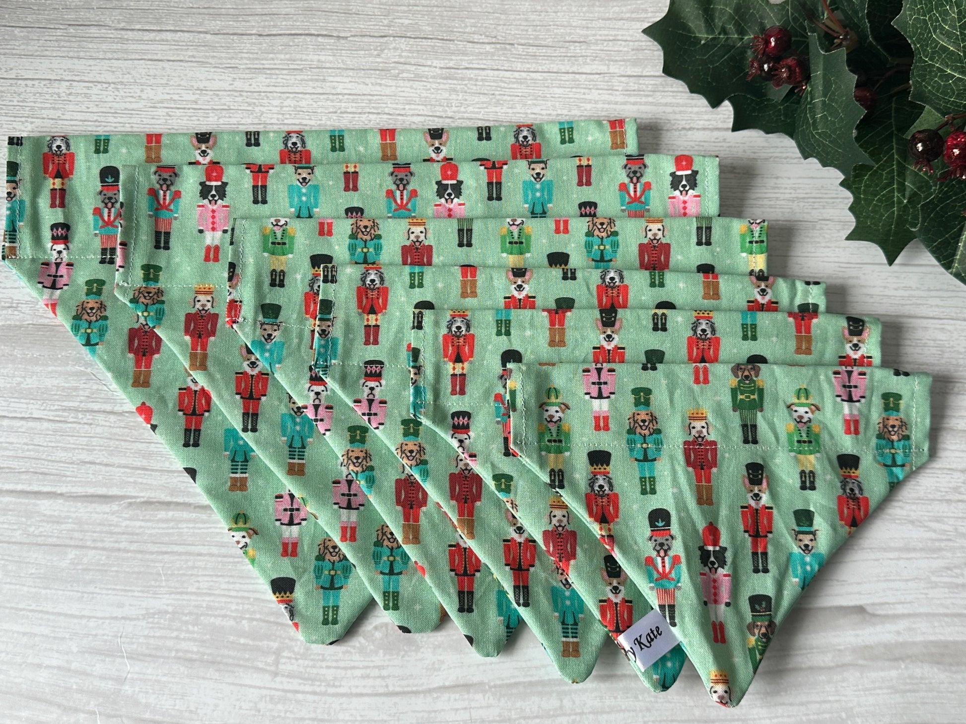 Five green cloth napkins with a nutcracker soldier pattern are fanned out on light wood. Holly and berries in the corner echo the festive spirit of the Crafts by Kate Cotton Pet Bandana with collar attachment—Puppy Nutcracker.