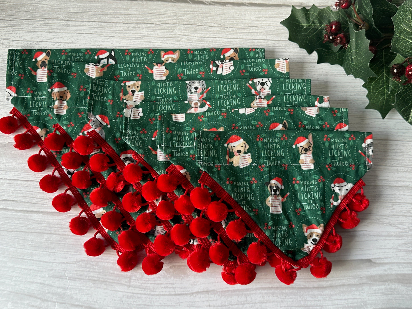 The "Licked it Twice" Cotton Pet Bandana by Crafts by Kate features cartoon dogs in holiday outfits and text on green cotton, finished with red pom-pom trim and decorative holly and berries. Includes a convenient collar attachment.
