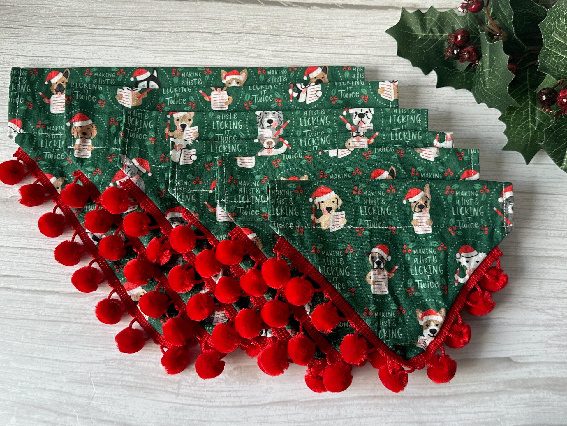 The "Licked it Twice" Cotton Pet Bandana by Crafts by Kate features cartoon dogs in holiday outfits and text on green cotton, finished with red pom-pom trim and decorative holly and berries. Includes a convenient collar attachment.
