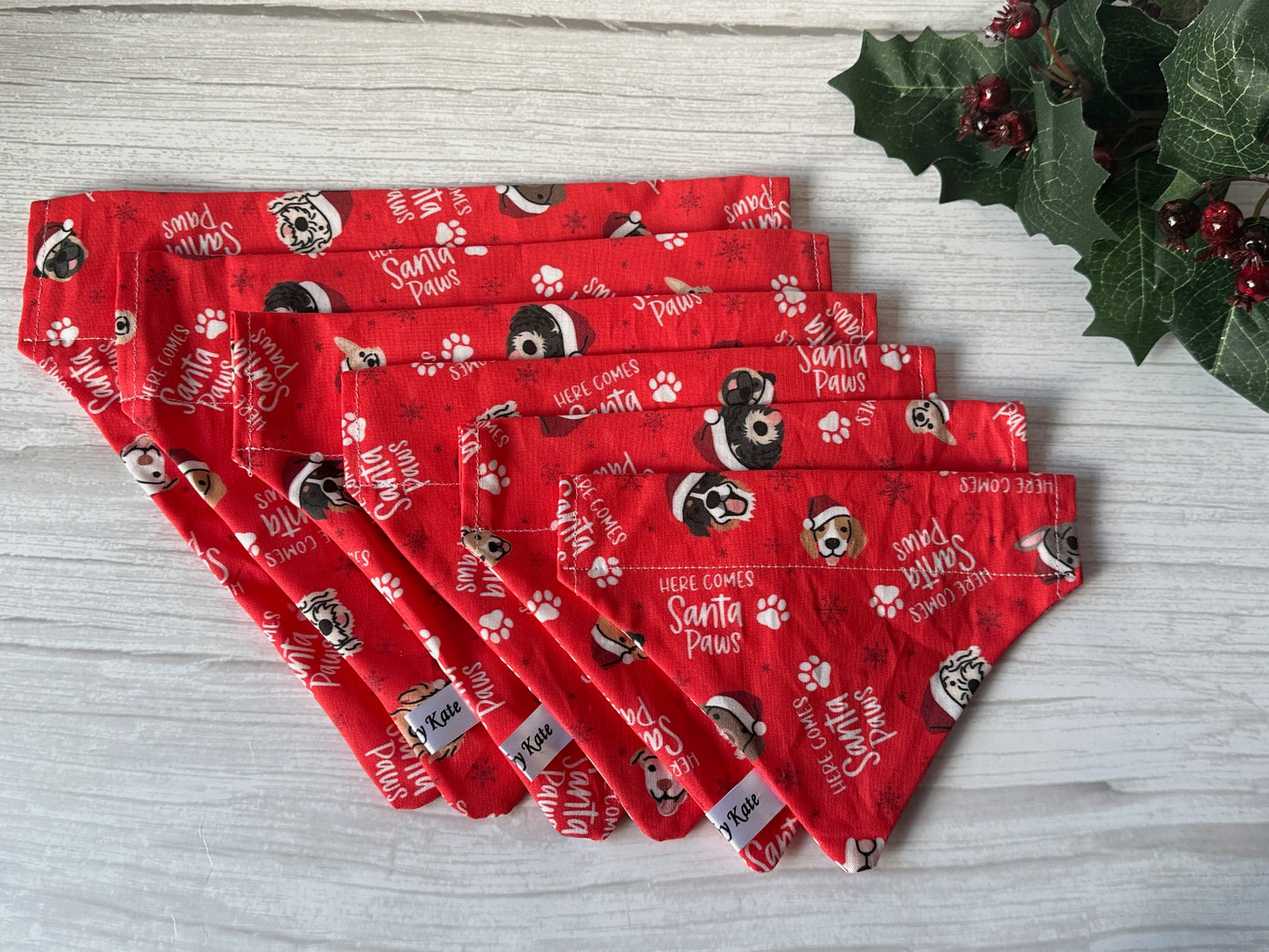 Six red "Here comes Santa Paws" cotton pet bandanas by Crafts by Kate, featuring playful holiday dog faces, paw prints, and holly accents, are fan-arranged on a white wooden background. Bandanas attach easily to your pet's collar.