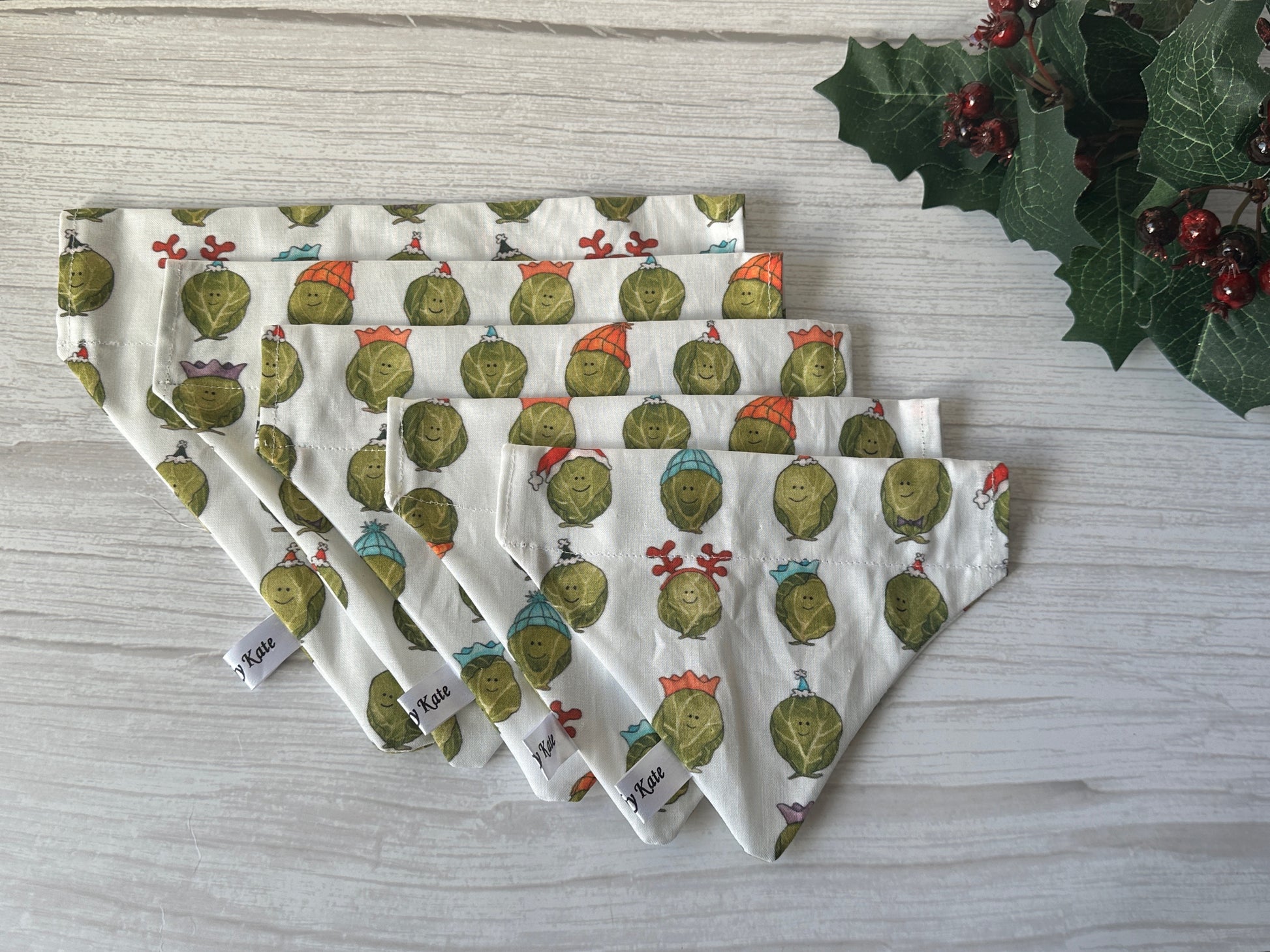 Five handcrafted "Party Sprouts" cotton pet bandanas by Crafts by Kate, each featuring smiling Brussels sprout faces in holiday hats, are displayed on a light wooden surface with holly leaves and berries for a festive touch.