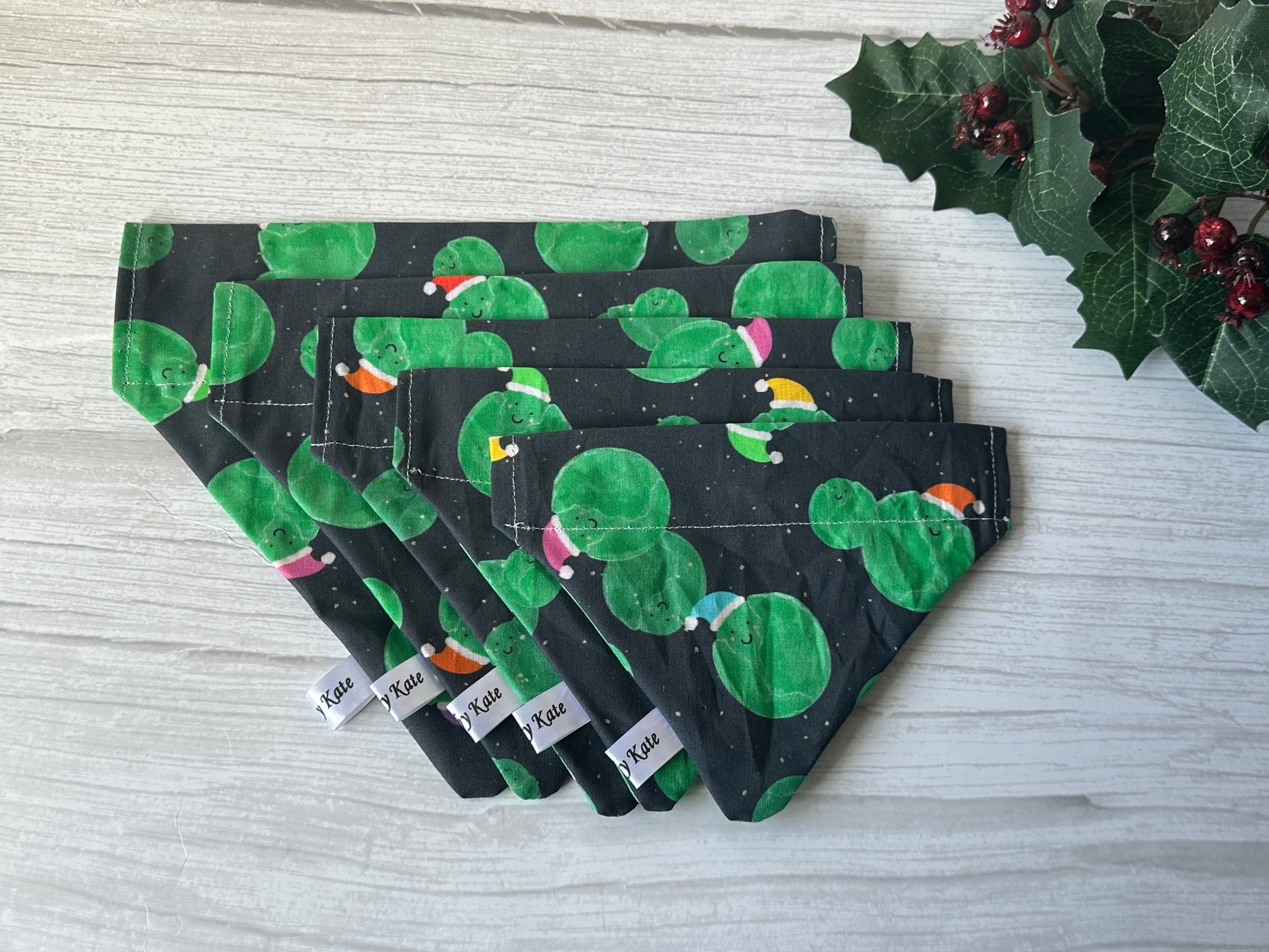 Five Crafts by Kate Cotton Pet Bandana with collar attachment - Santa Sprouts, featuring black fabric with green cactus and colorful bird patterns, are stacked on a light wood surface. A holly sprig with red berries is in the upper right corner.