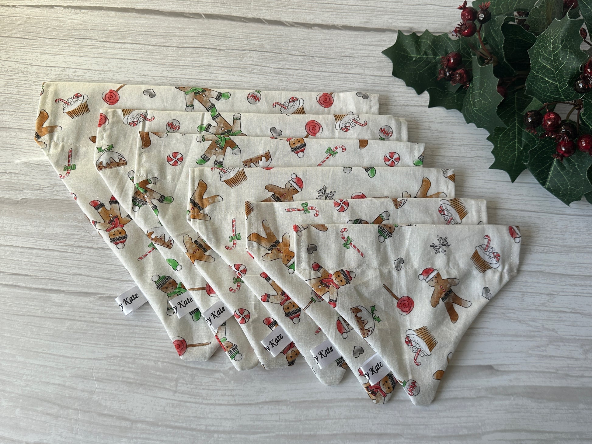 Six festive Crafts by Kate Cotton Pet Bandanas with collar attachment in Gingerbread Bakers fabric are fanned on light wood, holly and red berries at top right. Each comes in graduated sizes for your pup.
