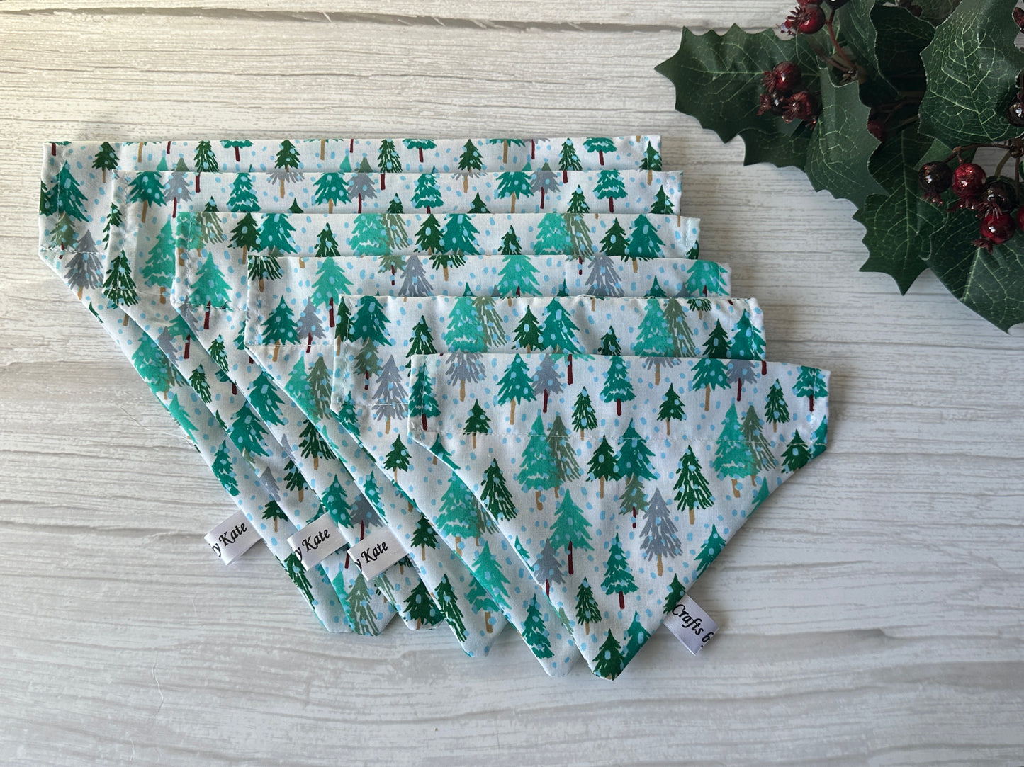 Five Crafts by Kate Cotton Pet Bandanas with collar attachment – Winter Forest, featuring green Christmas trees, are fan-arranged on light wood. A festive holly with red berries decorates the top right corner.