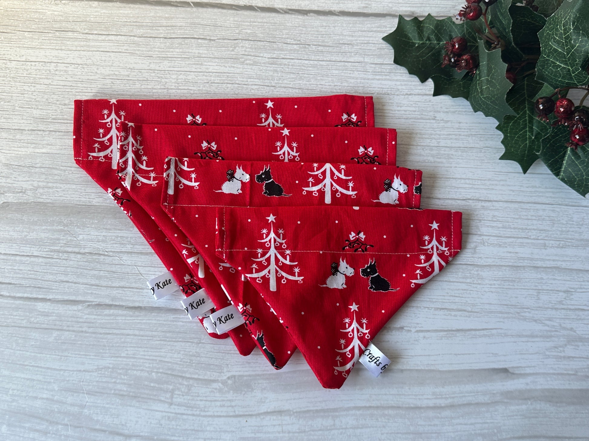 Four red "Cotton Pet Bandana with collar attachment - Festive Scottish Terrier" by Crafts by Kate are displayed on a white wooden surface, featuring white trees and Scottie dog prints with holly leaves and berries adding a festive touch.