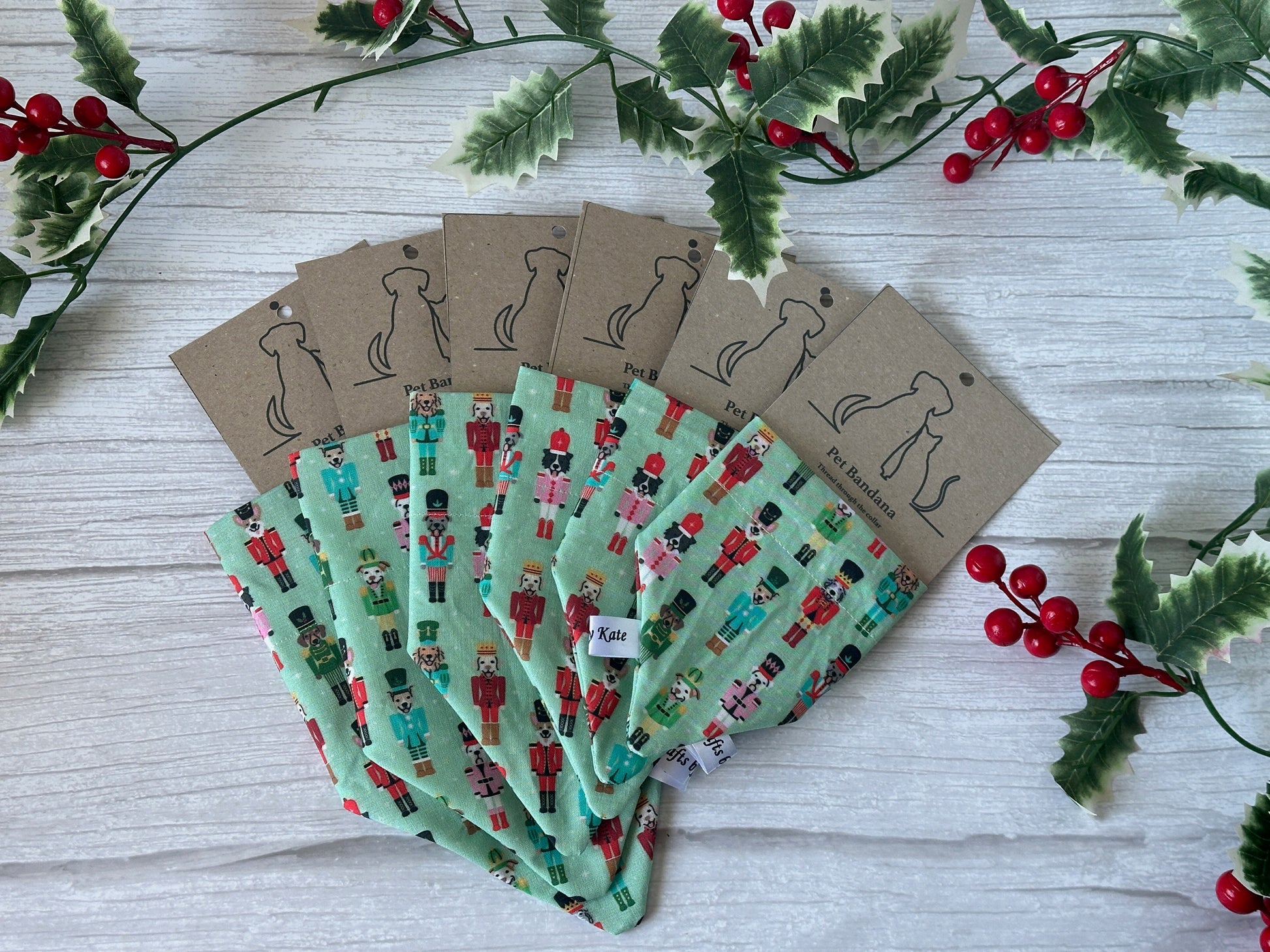 Six “Cotton Pet Bandana with collar attachment - Puppy Nutcracker” by Crafts by Kate are fanned out on a white surface, each on a dog-logo card. Holly leaves and berries accent this festive display of cotton pet bandanas.