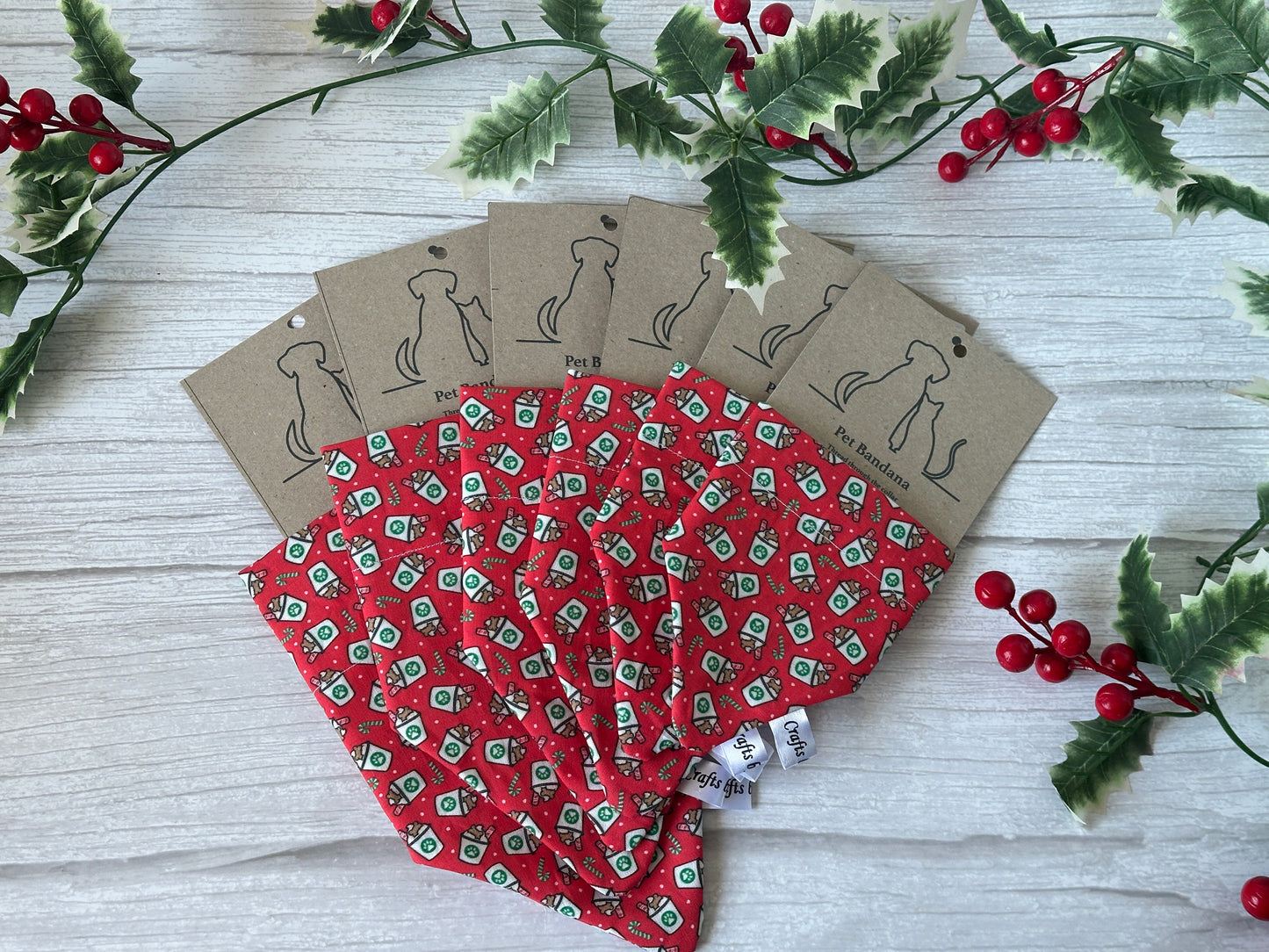 Six Crafts by Kate Cotton Pet Bandanas with collar attachments, featuring festive Christmas Pupcup patterns, are fanned out on a white wooden surface with holly and berry decor. Each has a brown tag with a dog outline—ideal for your dog's holiday celebration.