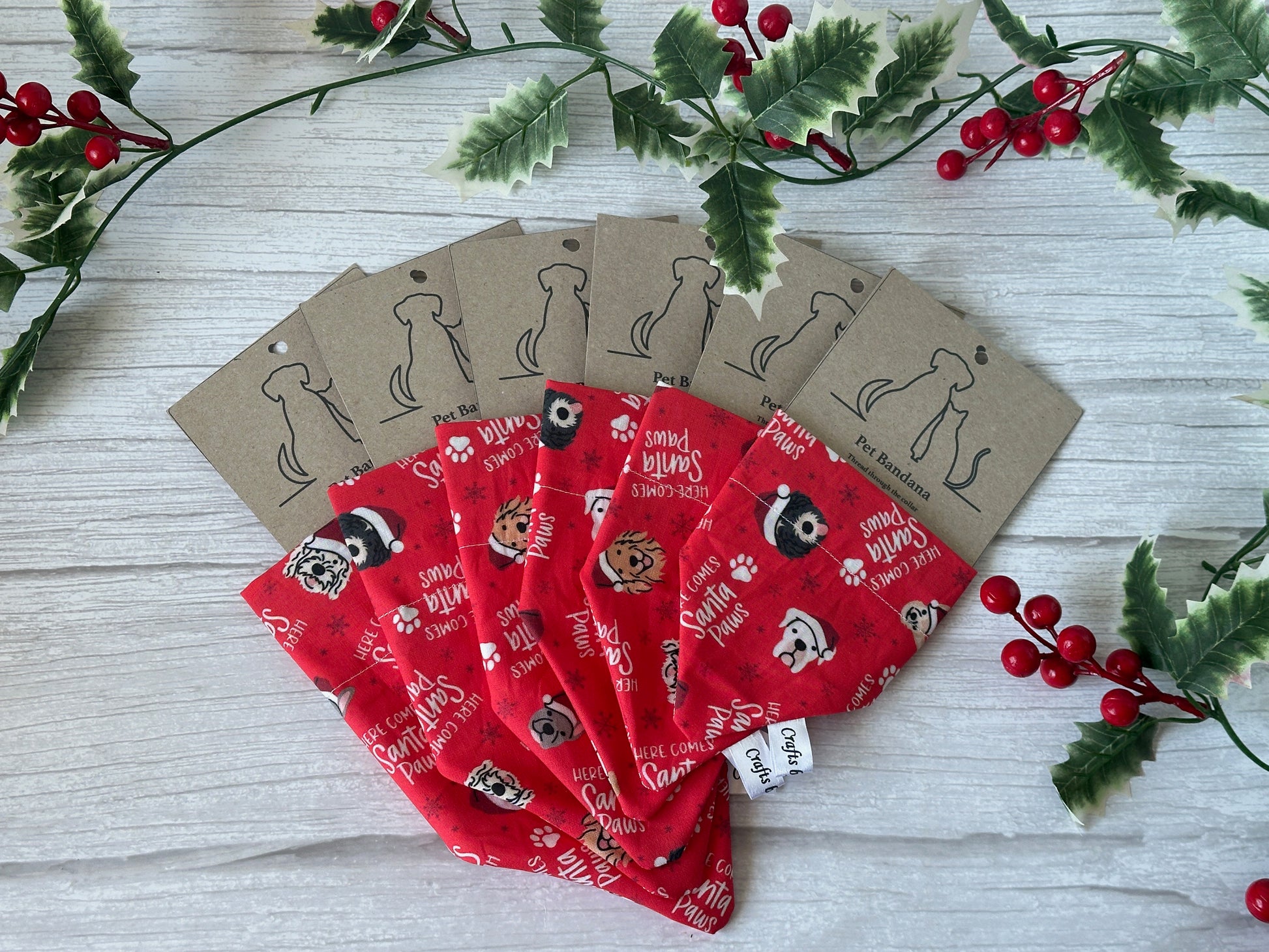 Six red "Here comes Santa Paws" cotton pet bandanas by Crafts by Kate are fanned out on a white wooden surface, surrounded by green holly and red berries. Each features a collar attachment and kraft tag with dog outline.