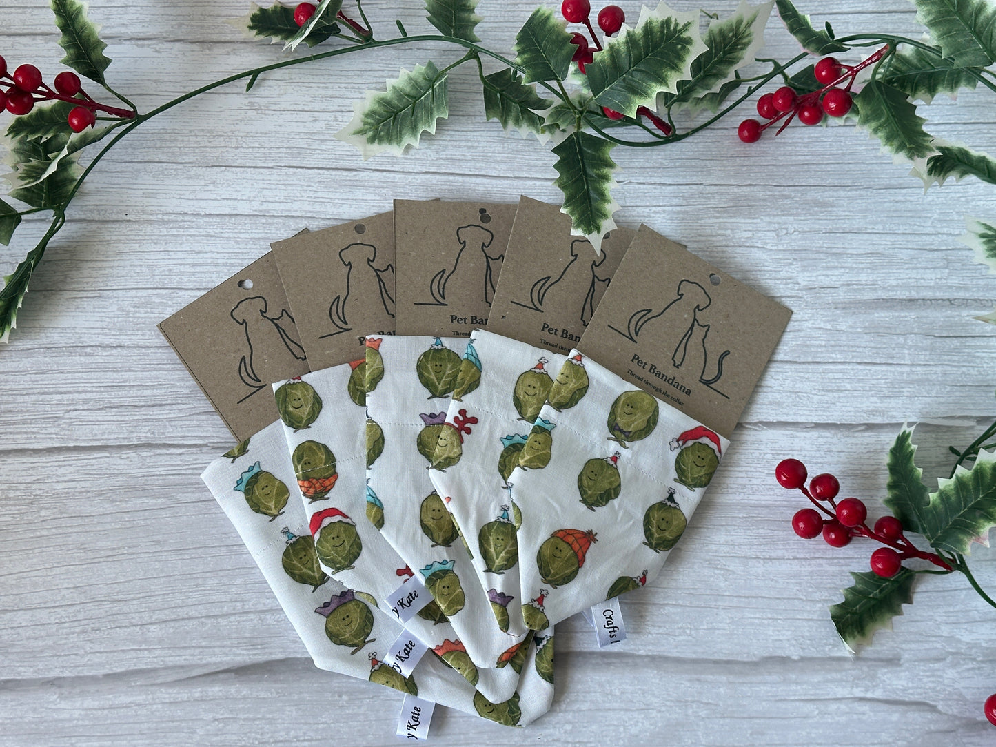 Five handcrafted "Party Sprouts" cotton pet bandanas by Crafts by Kate, each featuring a collar attachment and brown tag with dog silhouettes, are fanned out on a white wooden background bordered by holly branches with red berries.