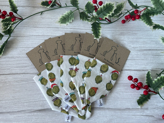 Five handcrafted "Party Sprouts" cotton pet bandanas by Crafts by Kate, each featuring a collar attachment and brown tag with dog silhouettes, are fanned out on a white wooden background bordered by holly branches with red berries.