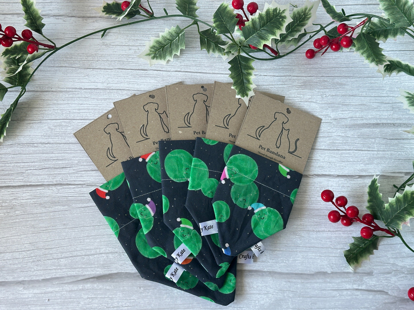 Five Crafts by Kate Cotton Pet Bandanas with collar attachment in the Santa Sprouts design, featuring green leafy patterns and brown dog silhouette tags, are displayed on a light wooden surface with holly sprigs.