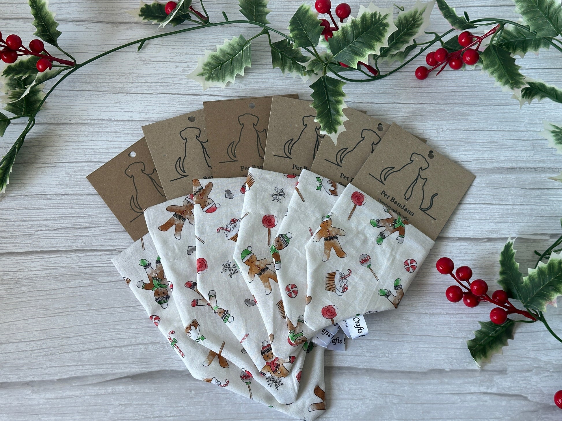 Five festive Cotton Pet Bandanas with collar attachment—Gingerbread Bakers by Crafts by Kate—featuring gingerbread and candy patterns are fanned out on light wood, surrounded by faux holly and brown dog-outline tags.