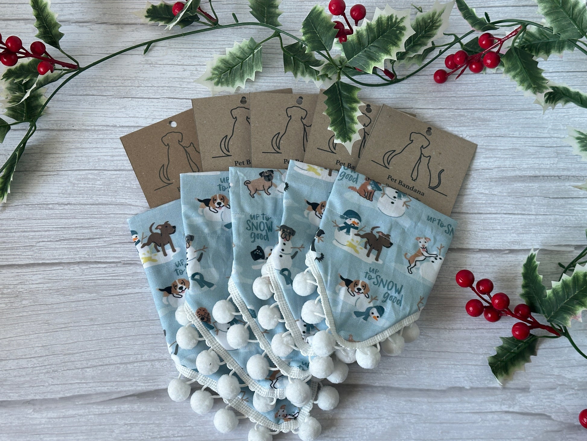 Five "Up to Snow-good" cotton pet bandanas by Crafts by Kate, each with paw and snowman prints and collar attachments, are arranged in a fan shape on a light wooden surface surrounded by holly and red berries.