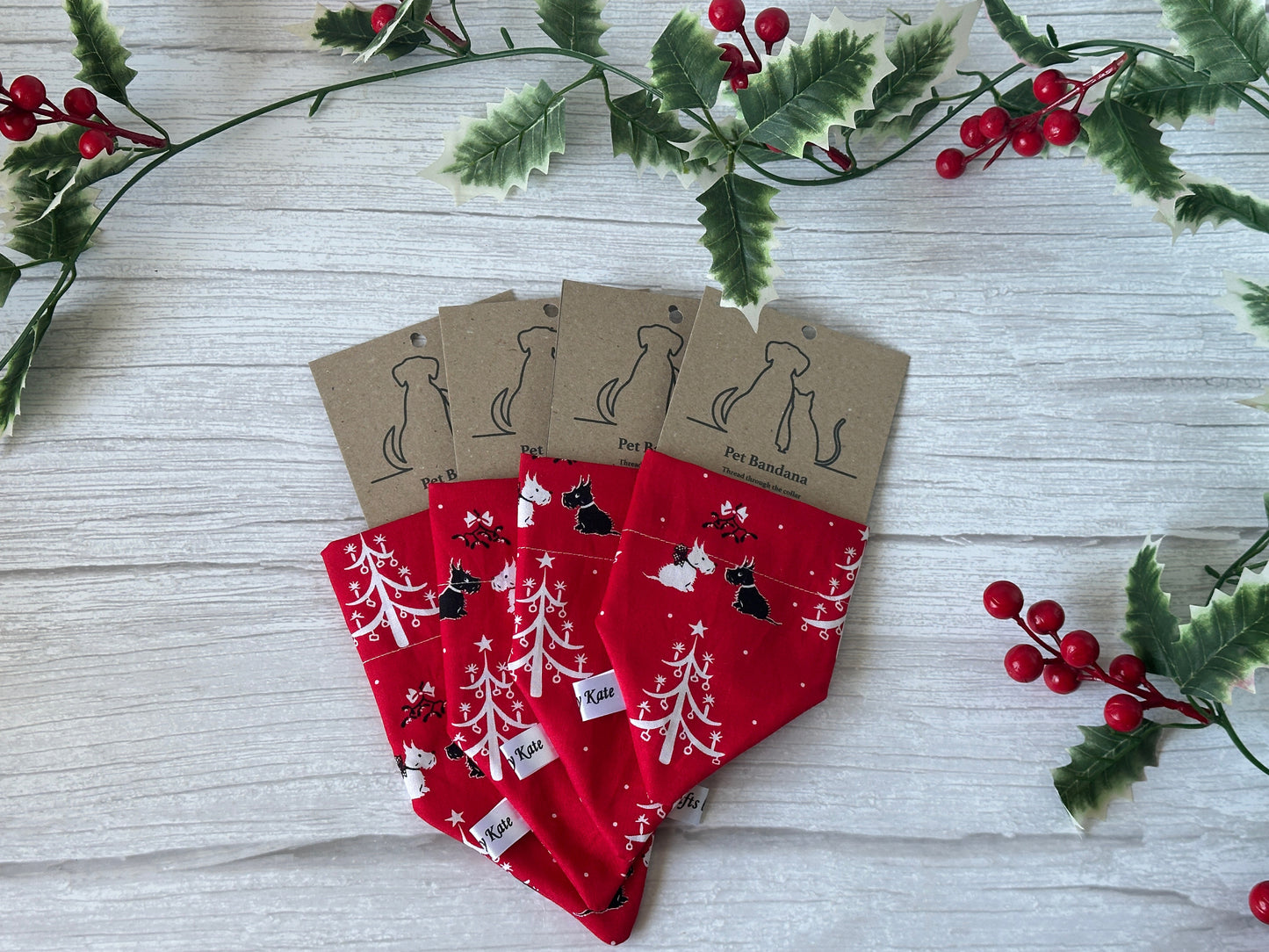 Four red Crafts by Kate cotton pet bandanas with collar attachment, featuring white Festive Scottish Terrier designs, are arranged on a light wood surface with holly branches. Each accessory has a brown tag with a pet silhouette illustration.