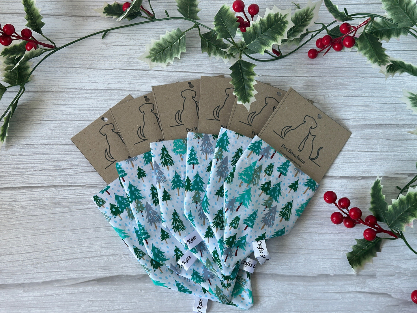 Five Cotton Pet Bandanas with collar attachment in the Winter Forest design by Crafts by Kate are arranged fanlike on a white wooden surface, decorated with holly leaves and red berries.