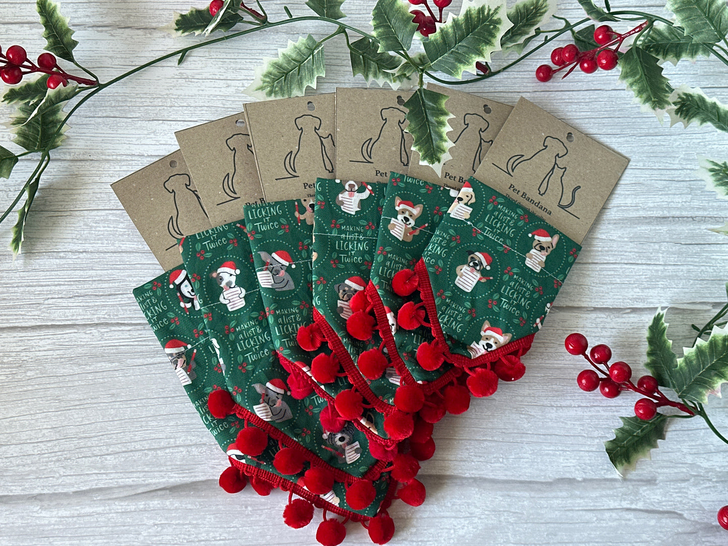 Five green "Licked it Twice" cotton pet bandanas by Crafts by Kate, each with red pom-pom trim, are fanned out. Every bandana features a collar attachment and hangs from a brown, holly-embellished tag with a dog silhouette.