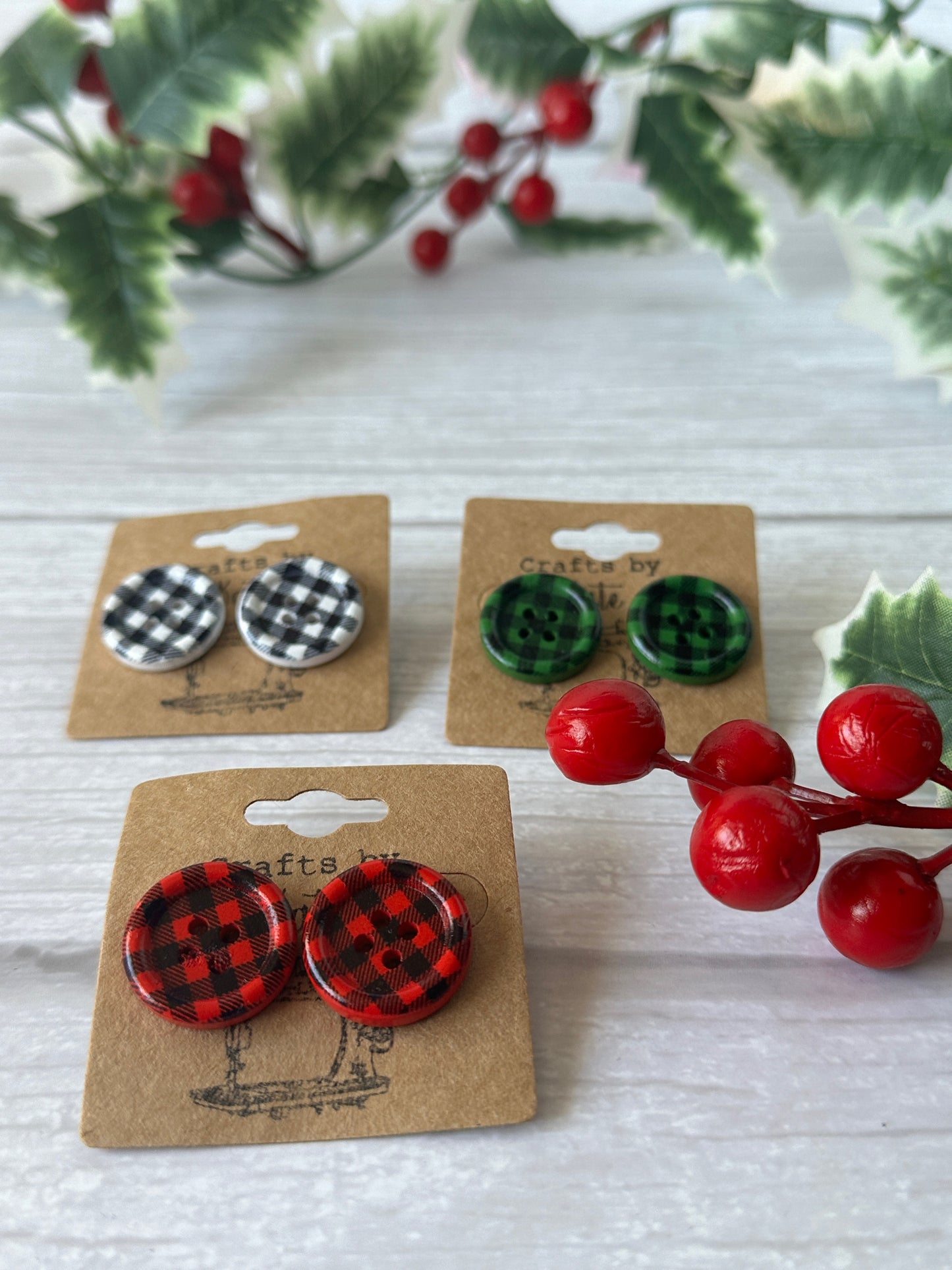 Three pairs of Crafts by Kate's Handcrafted Button Tartan Stud Earrings in red, green, and black-and-white are displayed on brown cards amidst festive red berries and green leaves on a light wooden surface.