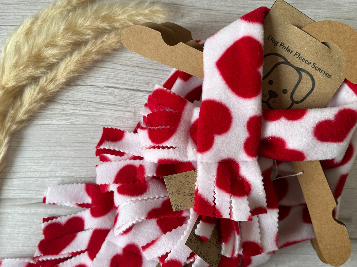 Crafts by Kate's Polar Cotton Fleece Pet Scarf - Red Hearts hangs elegantly on cardboard hangers. Each is displayed on a light wooden surface, complemented by a dried wheat stalk, and offers style and comfort for dogs.
