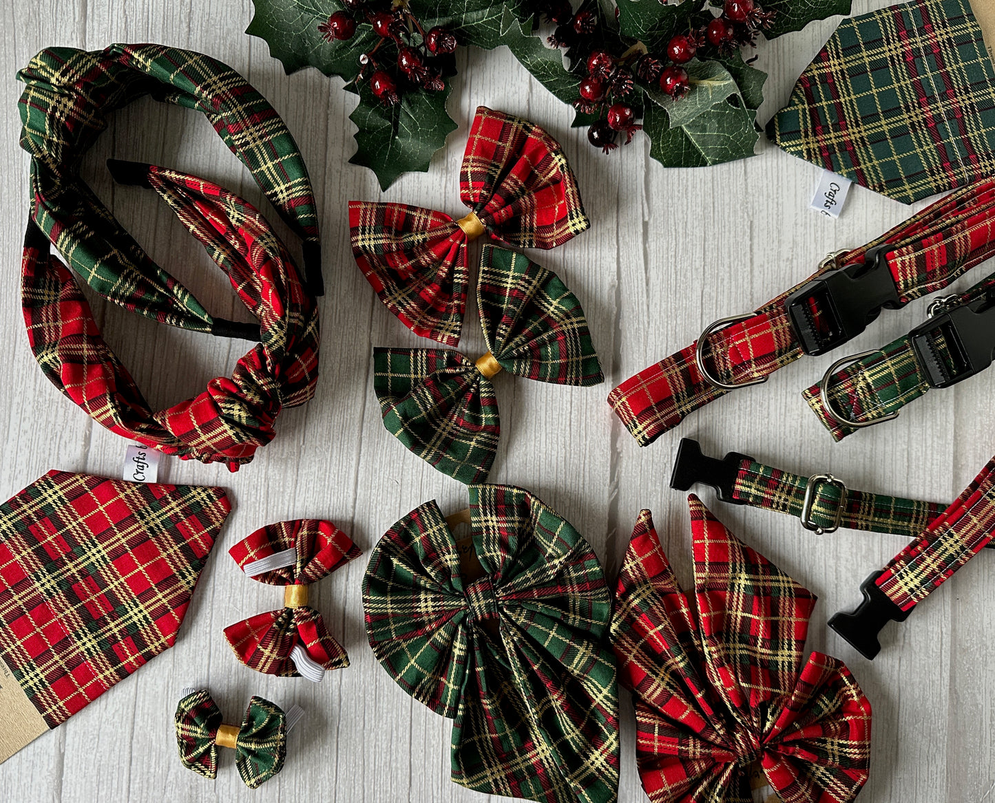 A festive flat lay showcases the Crafts by Kate Cotton Pet Bandana with collar attachment in Red Christmas Tartan, displayed on light wood with holly and berries, alongside matching bows, collars, headbands, and bow ties for a holiday vibe.
