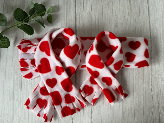 The Polar Cotton Fleece Pet Scarf - Red Hearts by Crafts by Kate is artfully displayed on light wood, highlighting the cozy charm of the material. A small green branch enhances the natural elegance of these handcrafted scarves.